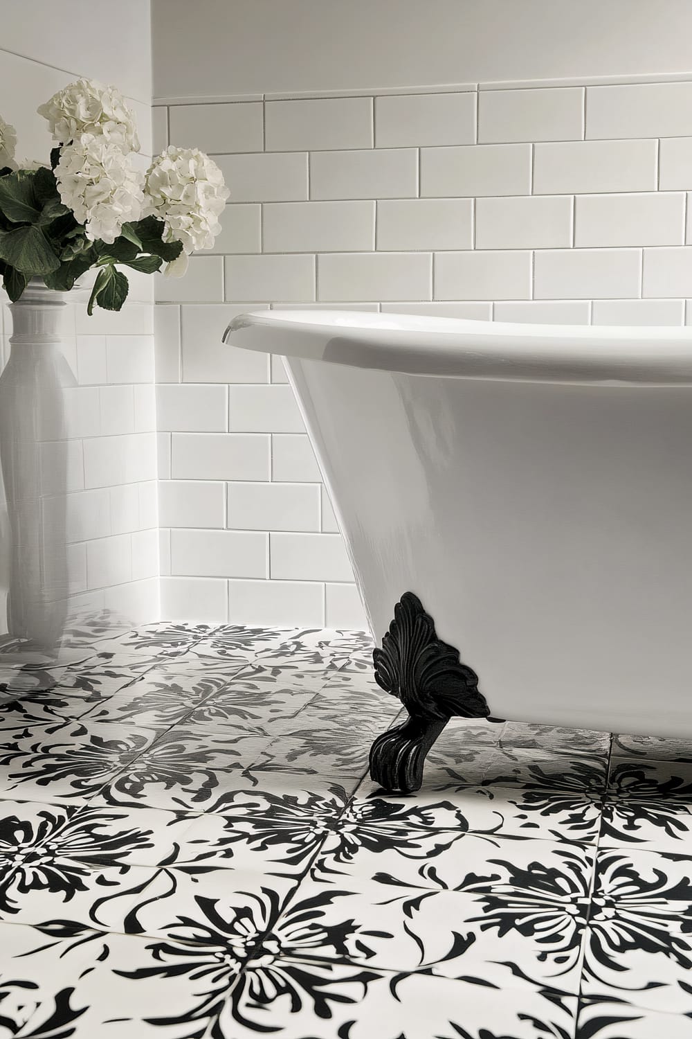 A Victorian-style bathroom corner showcasing a pristine clawfoot bathtub with black ornamental legs. The tub is set against a background of glossy white subway tiles. The floor features bold black-and-white patterned tiles in a floral design. A white vase filled with white hydrangeas sits on a ledge to the left of the tub.