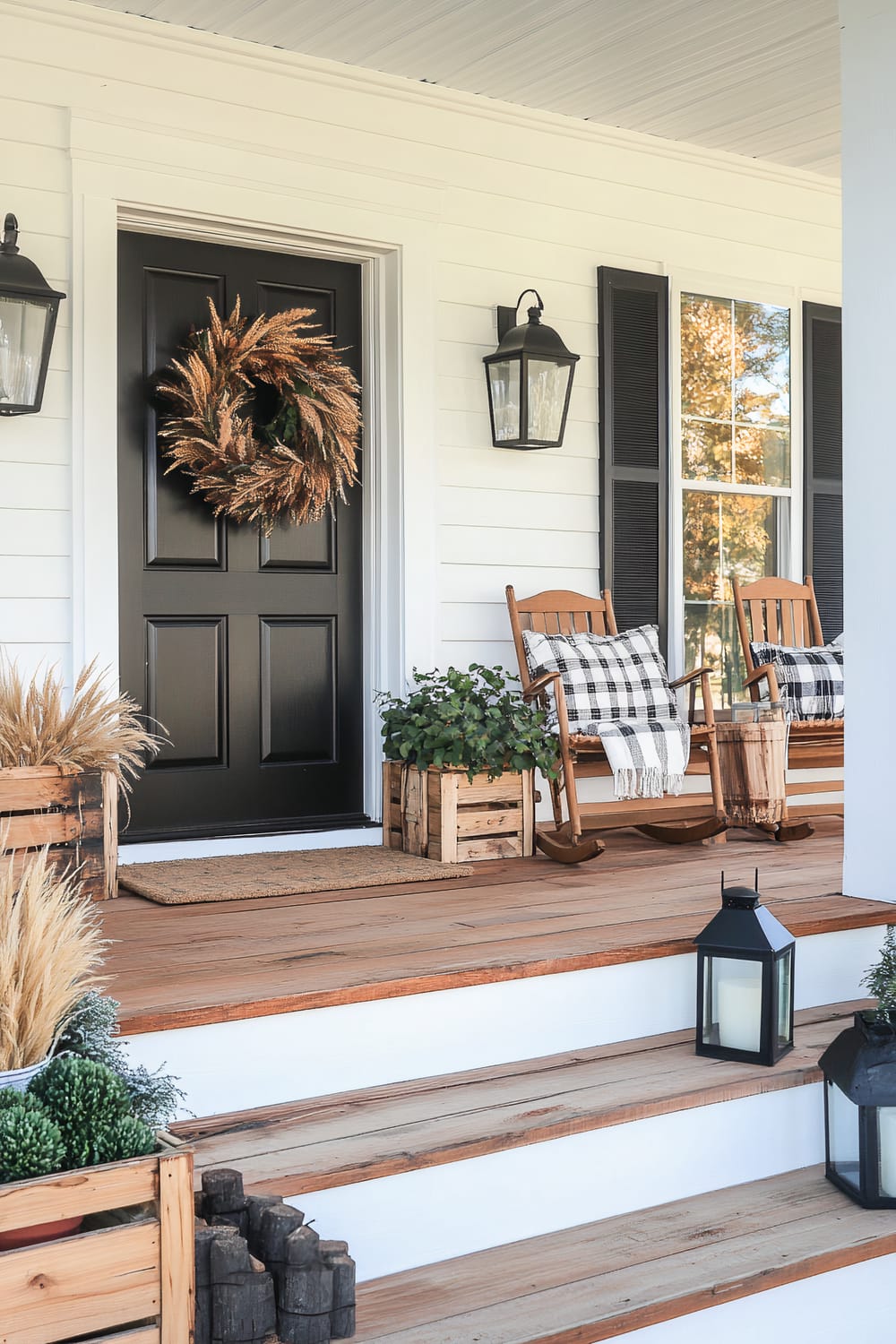 An inviting porch features a black front door adorned with a natural autumnal wreath. Black lantern-style wall sconces flank the door, providing ambient lighting. Two wooden rocking chairs with black and white plaid cushions and throws sit on either side, accompanied by wooden crates containing decorative plants. A natural fiber doormat rests in front of the door, and planters with seasonal foliage are placed along the wooden steps, which also host black metal lanterns with white candles inside.