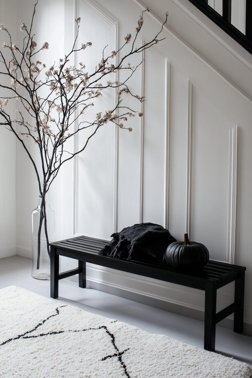 An image showcasing a minimalist interior with a black wooden bench accessorized with a black throw and a black pumpkin. The bench is set against a white paneled wall with a large glass vase containing tall, branchlike twigs with dried flowers. The floor features a light-colored rug with a simple black geometric pattern.