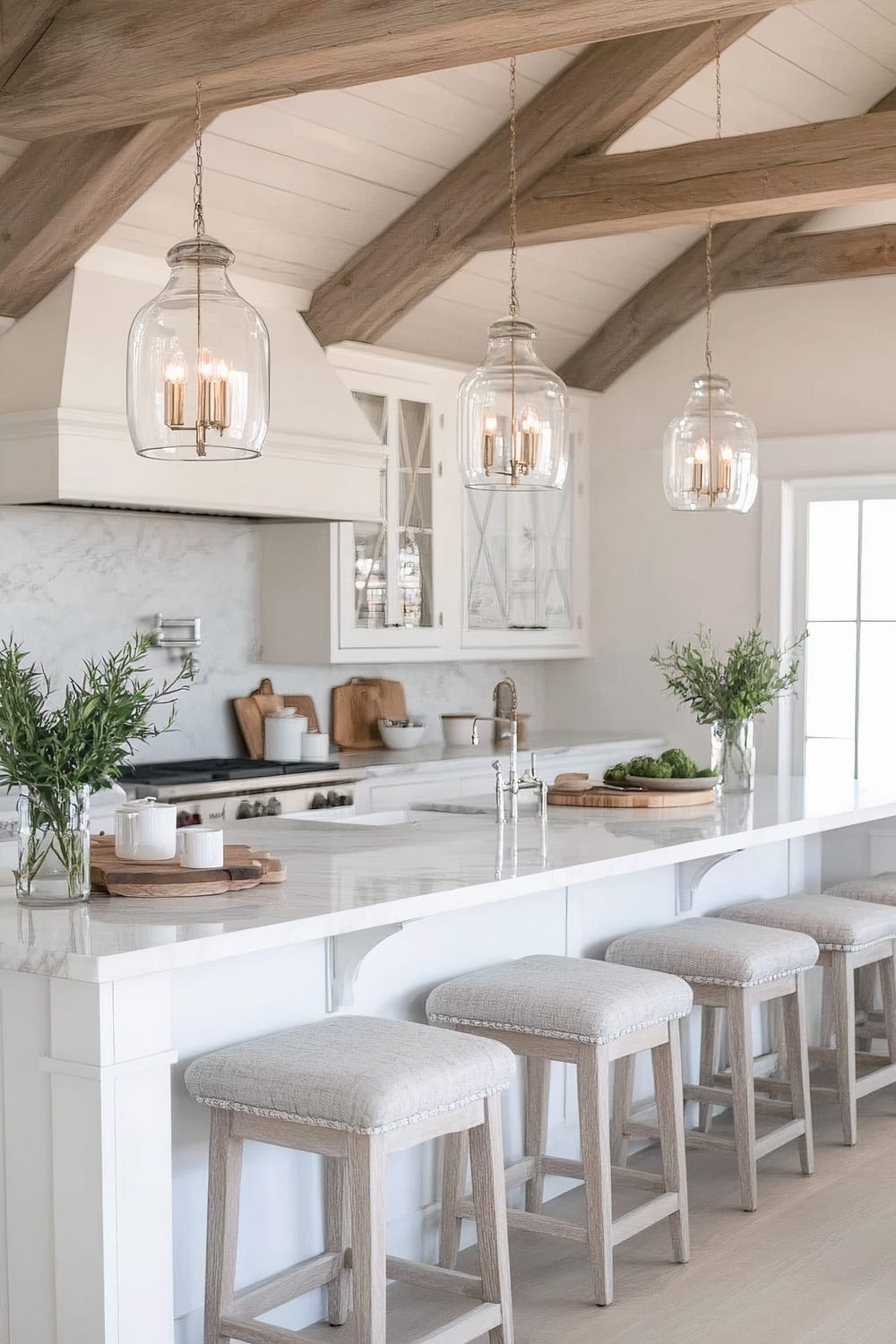 This image depicts a bright, modern kitchen. It features a long, white marble island countertop with five cushioned bar stools beneath it. Above the island hang three glass pendant lights with brass fixtures. Behind the island, the kitchen has white cabinetry, a matching white range hood, and a white marble backsplash. There are greenery arrangements in glass vases on the island and countertops, adding a touch of nature. The ceiling has exposed wooden beams, and natural light fills the space from a large window.