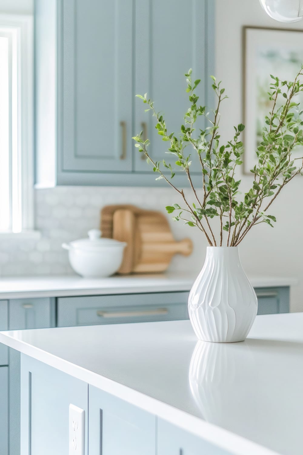 Close-up view of a kitchen corner showcasing light blue cabinets with a white countertop. The countertop holds a decorative white vase with green leafy branches. Wooden chopping boards and a white ceramic pot are also visible in the background. A window lets in natural light, illuminating the space.