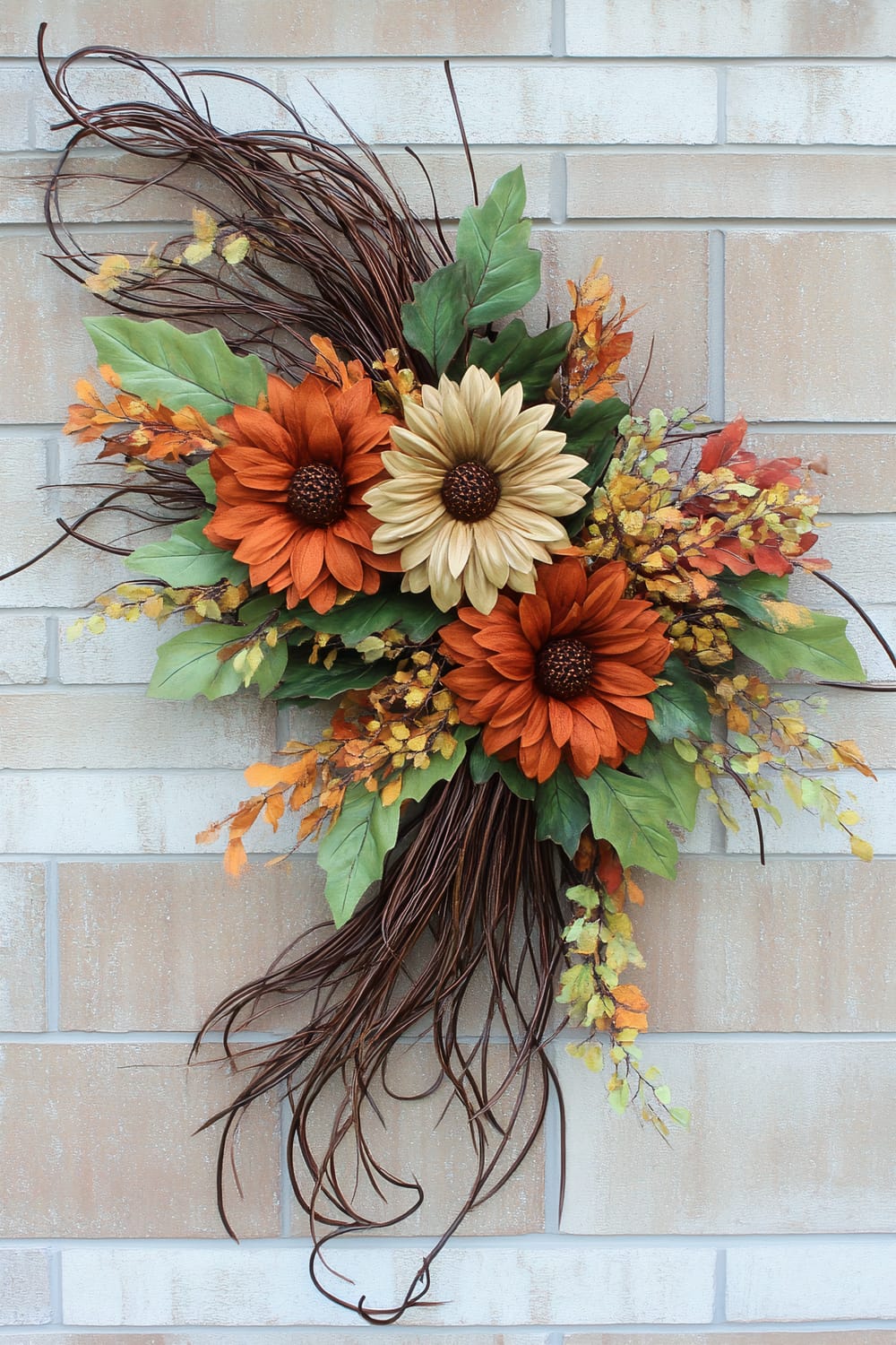 A vibrant autumnal wall decoration featuring a combination of artificial flowers and foliage on a neutral brick wall background. The arrangement includes two large orange flowers and one large beige flower at the center, surrounded by green leaves and smaller orange and yellow sprigs. Intertwined brown twigs extend outward, enhancing the naturalistic appearance.