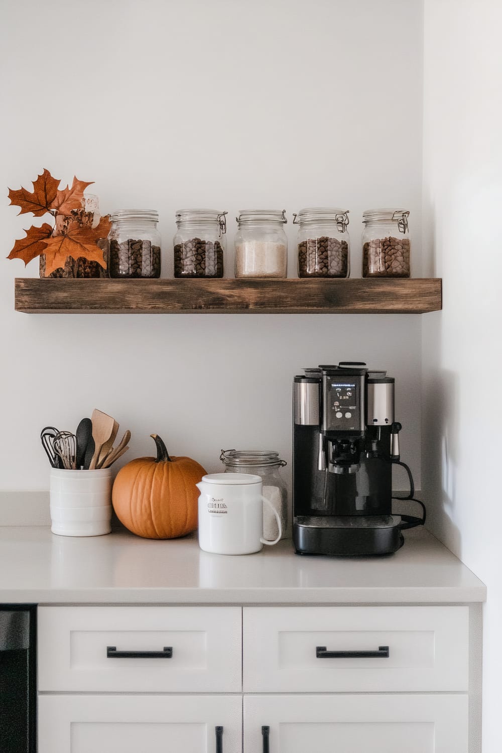 A kitchen countertop with a modern coffee machine on the right, a white ceramic mug, a glass jar filled with a white substance, and a pumpkin. On the left, a white crock holds various kitchen utensils, and above, a wooden floating shelf displaying jars of coffee beans and other dry goods with a decorative autumn leaf arrangement.