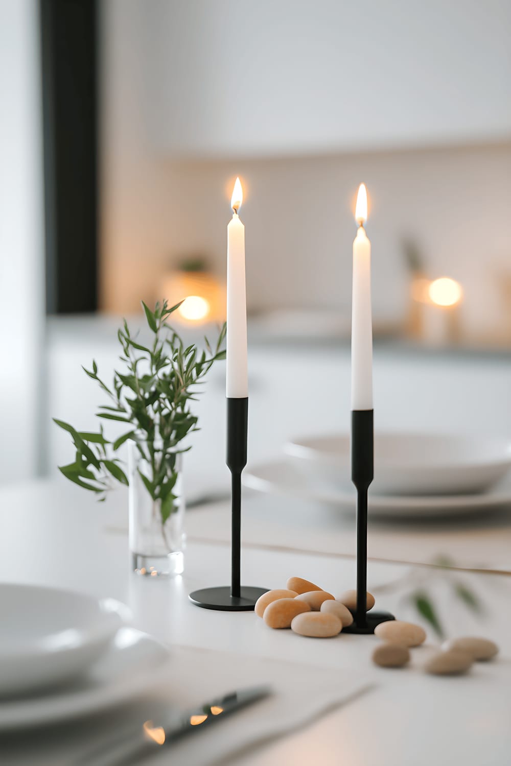 A minimalist centerpiece on a white table featuring pewter candle holders surrounded by small, soft peach-colored stones and green sprigs. The environment is Scandinavian in design with a bright, fresh, clean lighting.