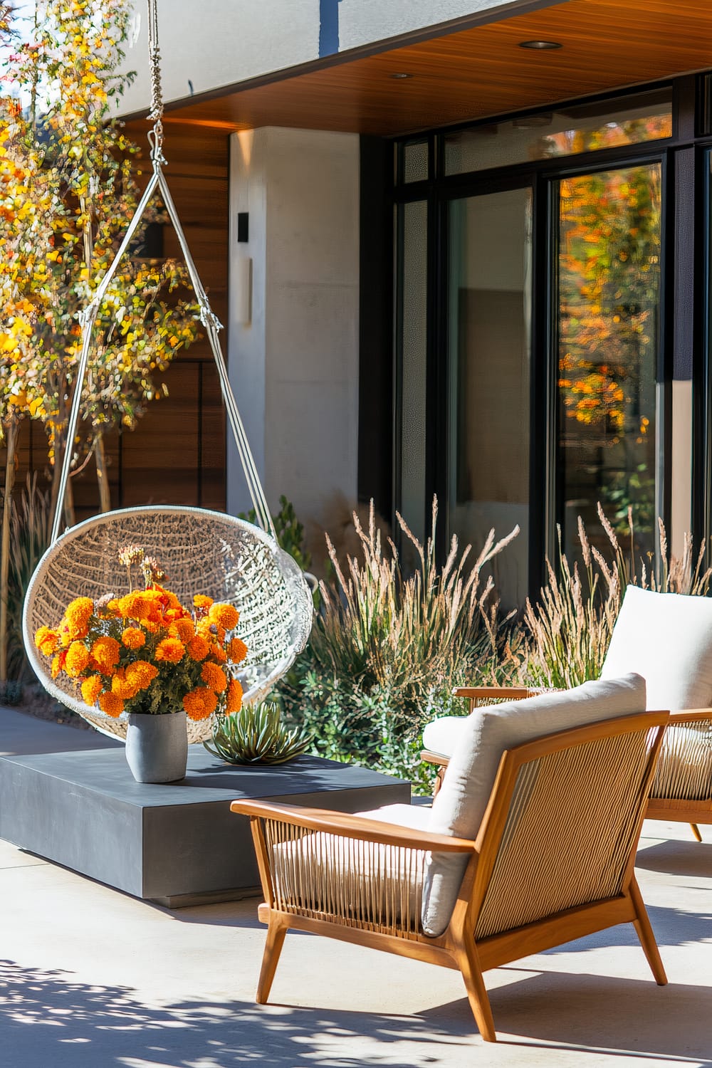 An outdoor seating area featuring a modern hanging chair with a macramé design, a concrete coffee table with a vase of bright orange flowers and a succulent, and an armchair with light-colored cushions. The background includes plants and a section of a house with wooden siding and large windows.