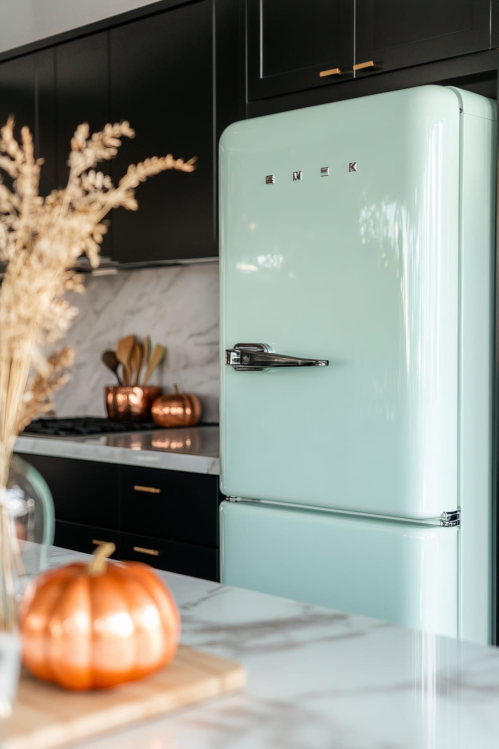 This image shows a kitchen with a light mint green retro-style refrigerator. The kitchen has black cabinetry with gold handles, and marble countertops. A copper pumpkin decoration and copper kitchen utensils are visible on the counter, along with a decorative vase with dried plants in the foreground.