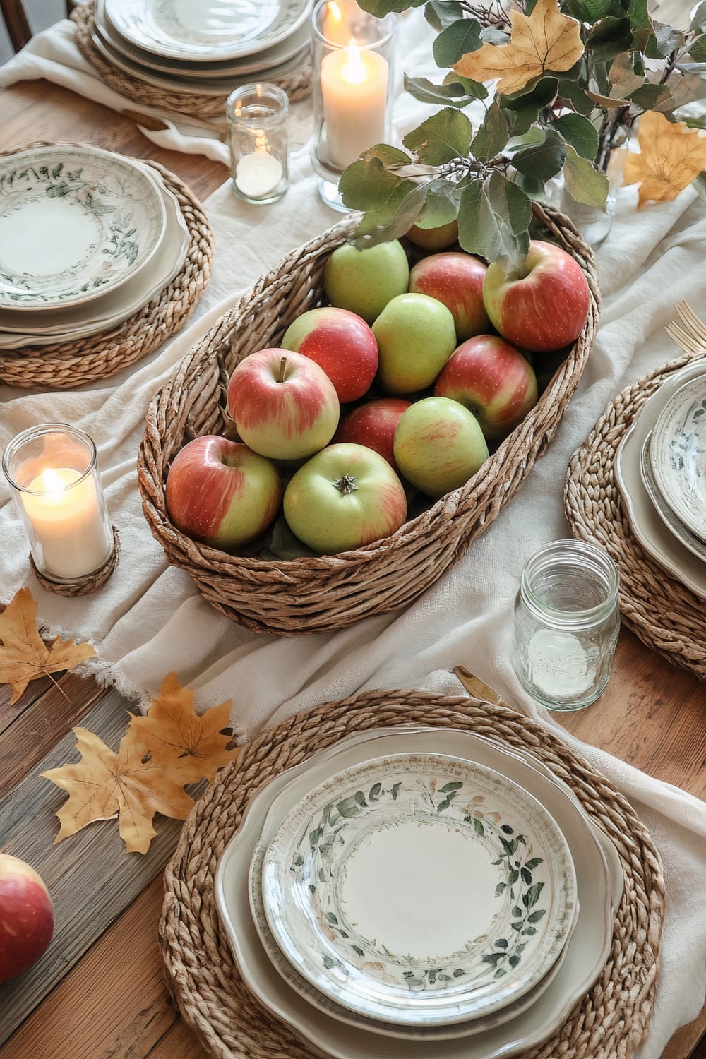 A rustic dining table setting arranged with a central woven basket filled with red and green apples. Surrounding the basket are candles in glass holders, some lit and others unlit, adding a warm glow to the scene. The table is set with woven placemats, each holding a set of stacked ceramic plates with a delicate floral pattern. A few autumn leaves are scattered around, and the table is draped with a light-colored cloth.