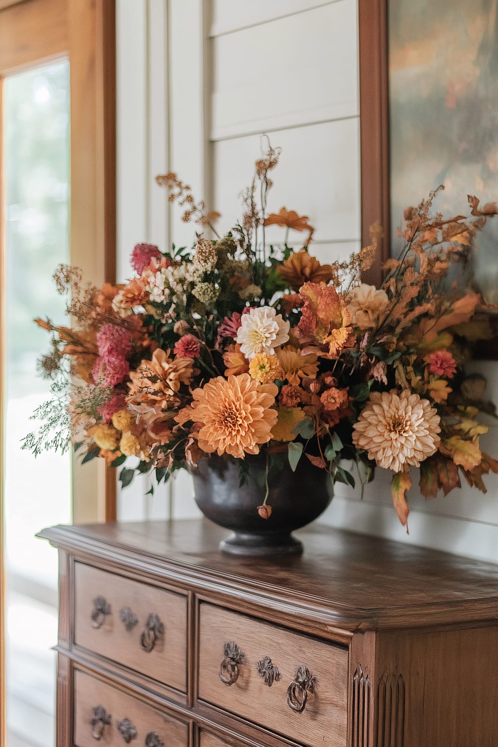 A dark, round vase overflowing with a vibrant array of autumn flowers sits atop a rustic, wooden dresser with intricately carved drawer handles. The flowers are in warm hues of orange, pink, and cream, complemented by dried foliage and greenery. The dresser is positioned against a white shiplap wall with a wooden-framed window to the left, allowing soft, natural light to illuminate the arrangement.