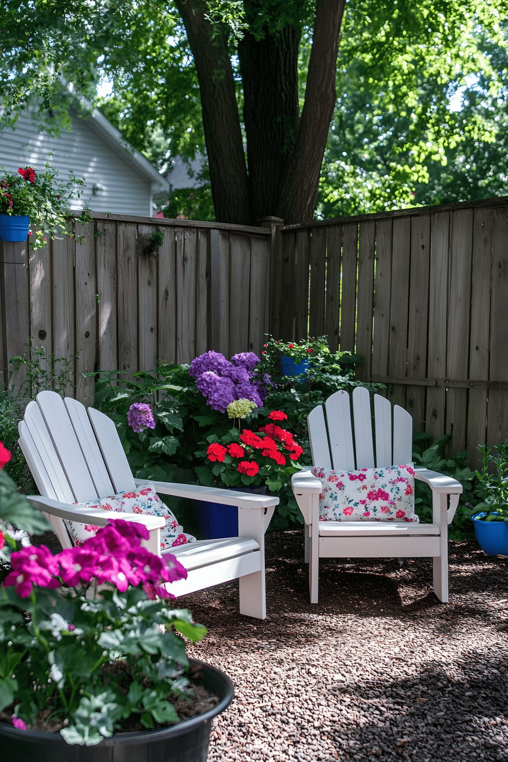 A budget-conscious backyard makeover embodying a seating area composed of two Adirondack chairs painted white, adjacent to a compact side table. The seating zone is romantically enveloped by lush green foliage and an array of vivid flowers housed in assorted pots and window boxes. A modest wooden fence, forming a natural backdrop, concludes the simple alfresco arrangement.