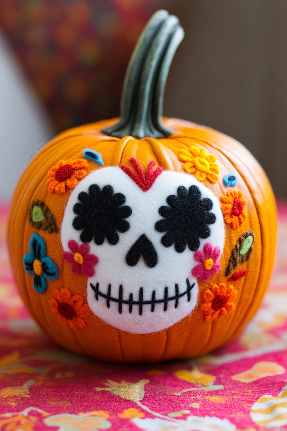 An orange pumpkin decorated with felt appliqués inspired by Day of the Dead motifs. The pumpkin features a white felt skull with black eyes and nose, and a stitched black mouth. Colorful felt flowers in red, blue, yellow, and pink surround the skull, creating a vibrant and festive appearance. The background is blurred but shows a colorful floral pattern, enhancing the festive feel.