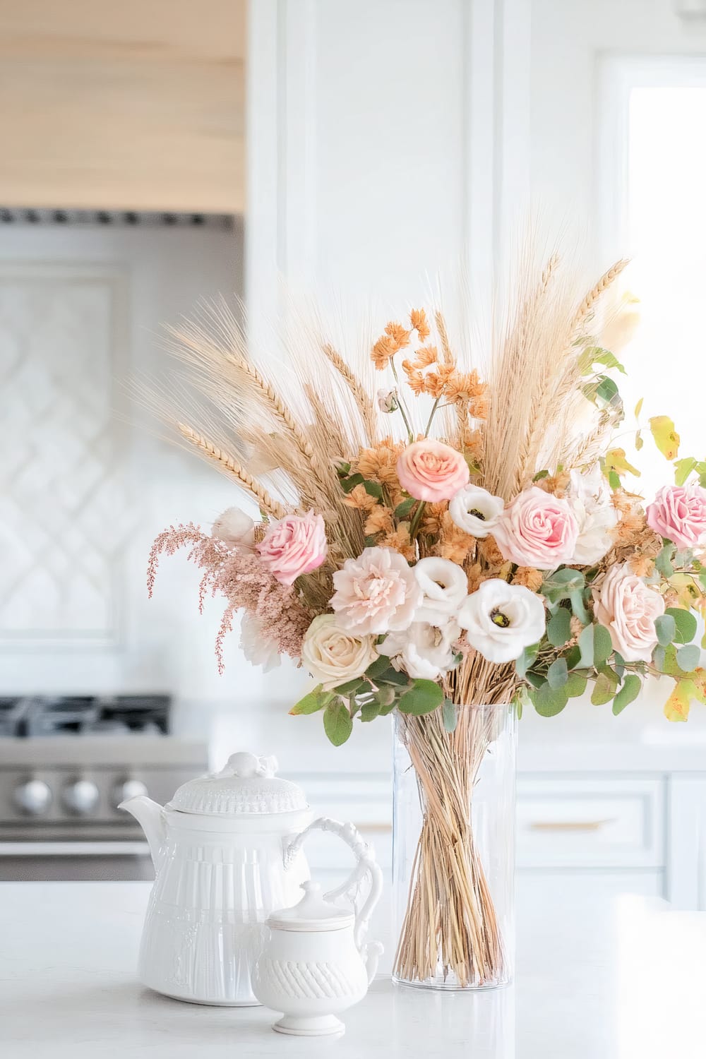 An elegant kitchen scene featuring a white countertop adorned with a clear glass vase filled with a mix of pastel-hued flowers, including pink and white roses, along with dried wheat stalks and yellow sprigs. Adjacent to the vase is a white ceramic teapot and a matching creamer set, adding a refined touch to the sophisticated setting. The background displays soft, neutral tones of cabinetry and a hint of a stovetop.