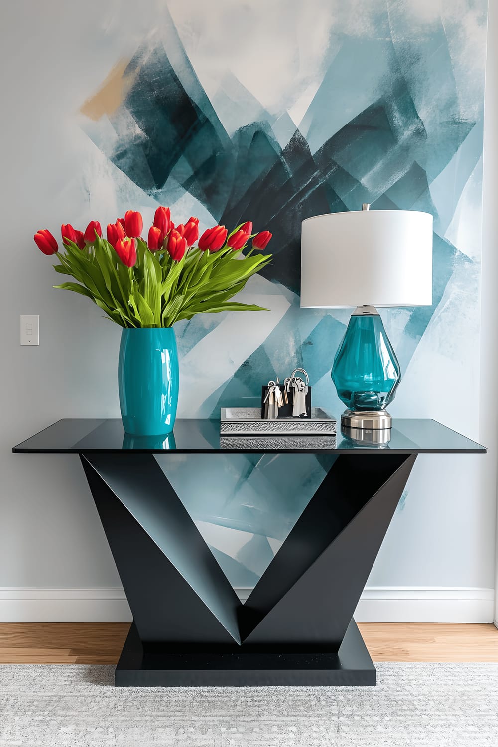 A stylish Milanese entryway featuring a bold geometric matte black console table with a glass top, adorned with a vibrant teal vase with bright tulips and a sleek silver tray with keys and a white-shaded lamp. A large abstract mural in teal, white, and gold shades is seen behind the console, while a grey runner covers the hardwood floor. A minimalist black coat rack with chrome hooks sits unobtrusively.