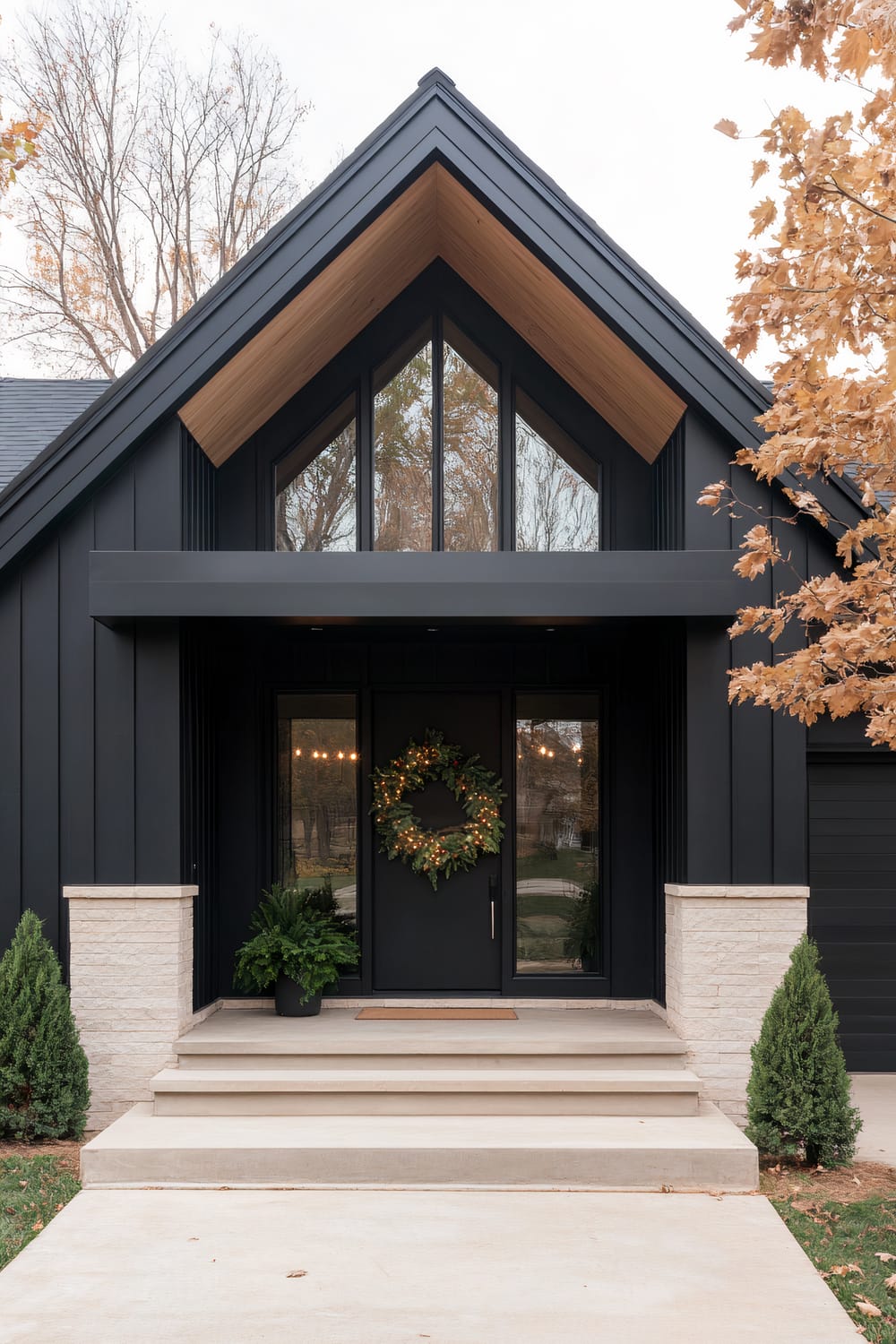 A modern home exterior display characterized by a steep, A-frame entryway with black vertical siding. A large, triangular window sits above the front door, allowing ample light to enter. The door is adorned with a holiday wreath, and plants are positioned on either side of the entry steps. The house features a blend of dark, sleek materials and lighter stone elements, with surrounding trees showing autumn foliage.