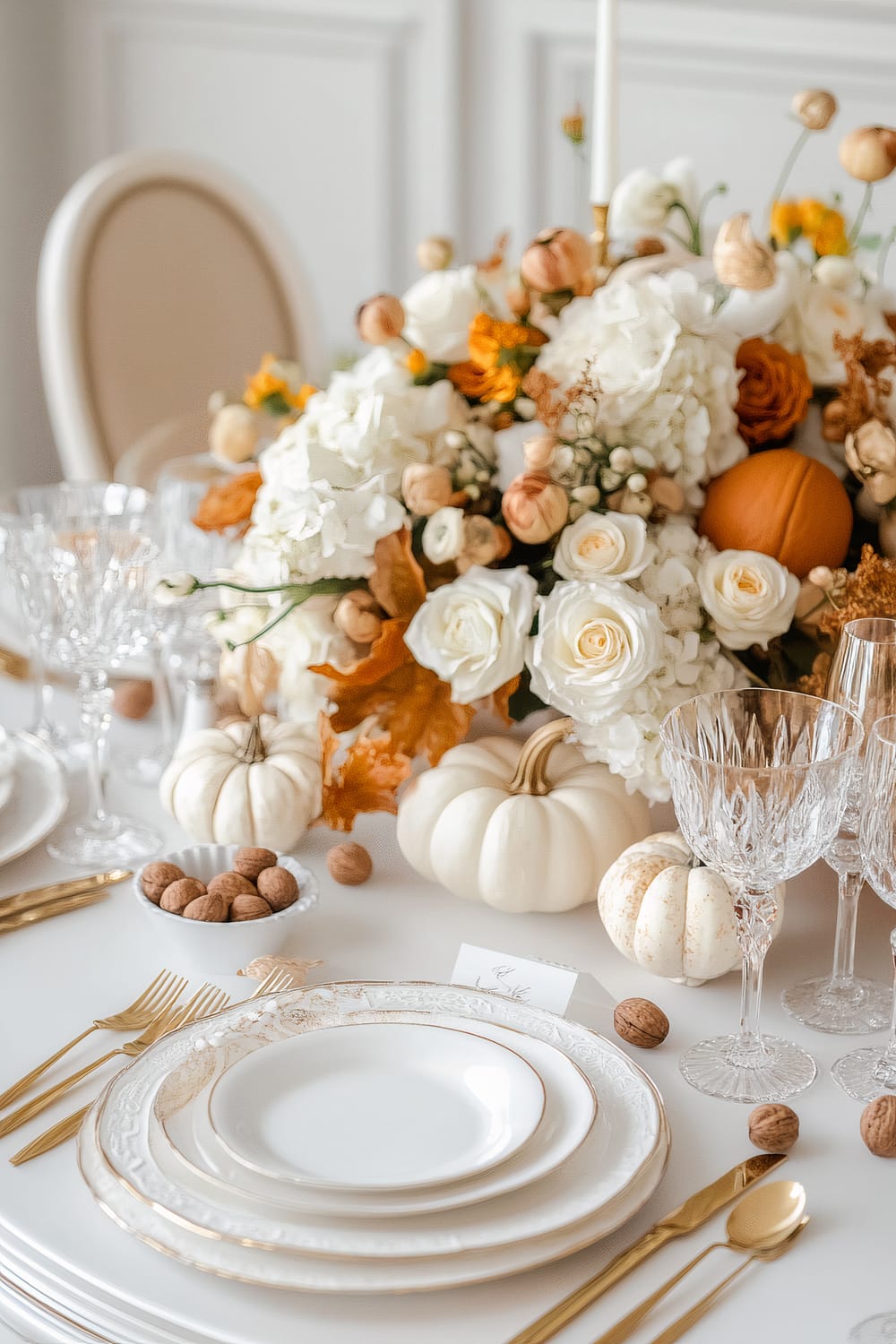 This image shows an elegant dining table set for a fall-themed gathering. The centerpiece is an opulent floral arrangement featuring white, cream, and orange flowers, complemented by small white and orange pumpkins. Crystal glassware and fine china with gold accents adorn the table, along with gold flatware meticulously arranged on the crisp white tablecloth. A bowl of walnuts and individual place cards add to the sophisticated ambiance.