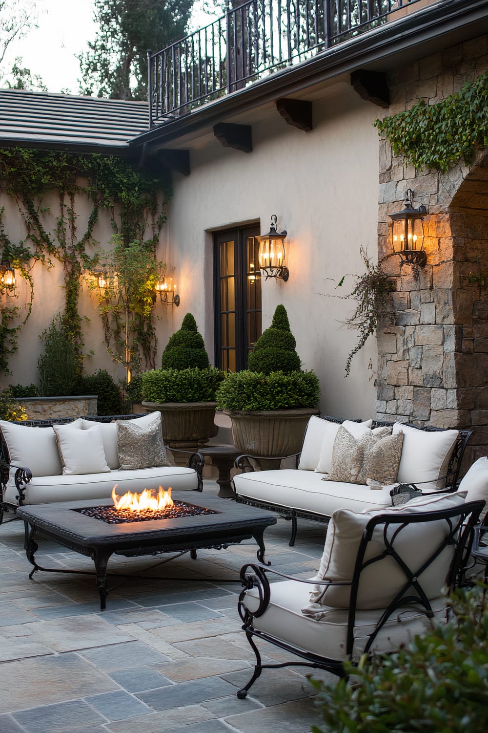 An elegant outdoor patio features wrought-iron furniture with white cushions arranged around a central fire pit table. The area is beautifully landscaped with potted greenery and climbing plants on the stucco walls. Stone cladding adds a rustic touch, complemented by wrought-iron wall-mounted lanterns that emit a warm glow.