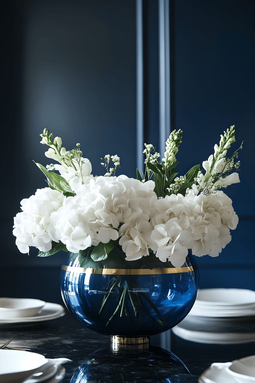 A stunning dining room scene with a brass floating flower arrangement in a cobalt blue glass bowl serving as the centerpiece on a sleek black marble table. The arrangement comprises white hydrangeas and green foliage, with brass accents intertwined among the flowers. The table is surrounded by minimalistic white dinnerware, and the whole scene is set against a backdrop of deep navy walls, gently illuminated by soft, ambient lighting.