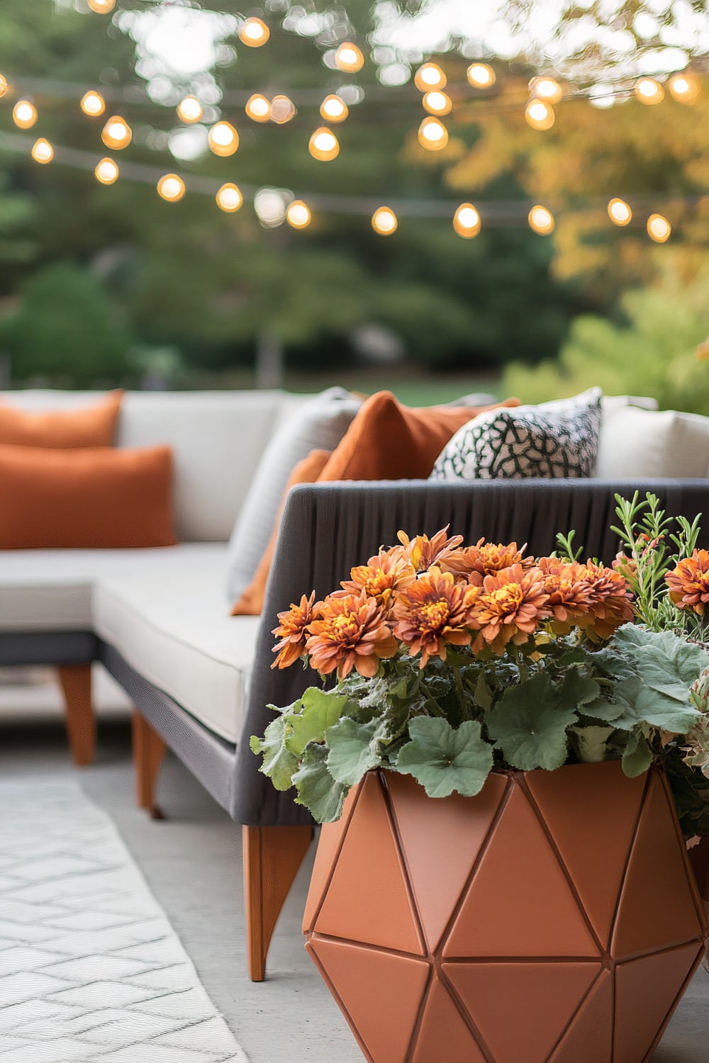 An outdoor patio scene showing a sectional sofa adorned with orange and patterned throw pillows. A geometric terracotta planter filled with orange flowers is in the foreground. String lights are hanging above, casting a warm glow, and a garden is visible in the blurred background.