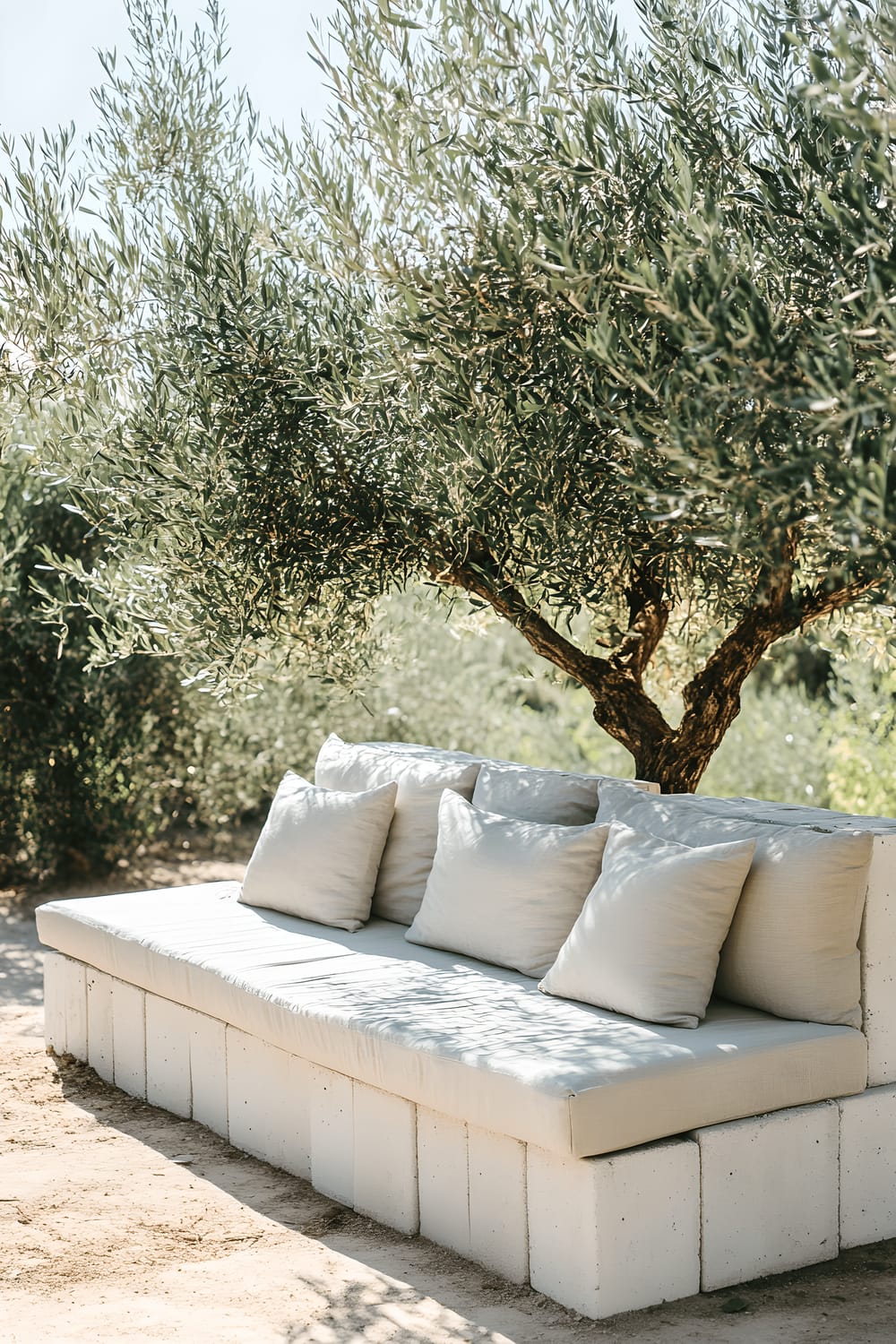 A minimalist outdoor setting featuring a homemade sofa made from whitewashed cinder blocks, topped with neutral linen cushions. The sofa is positioned outdoors, surrounded by olive trees.