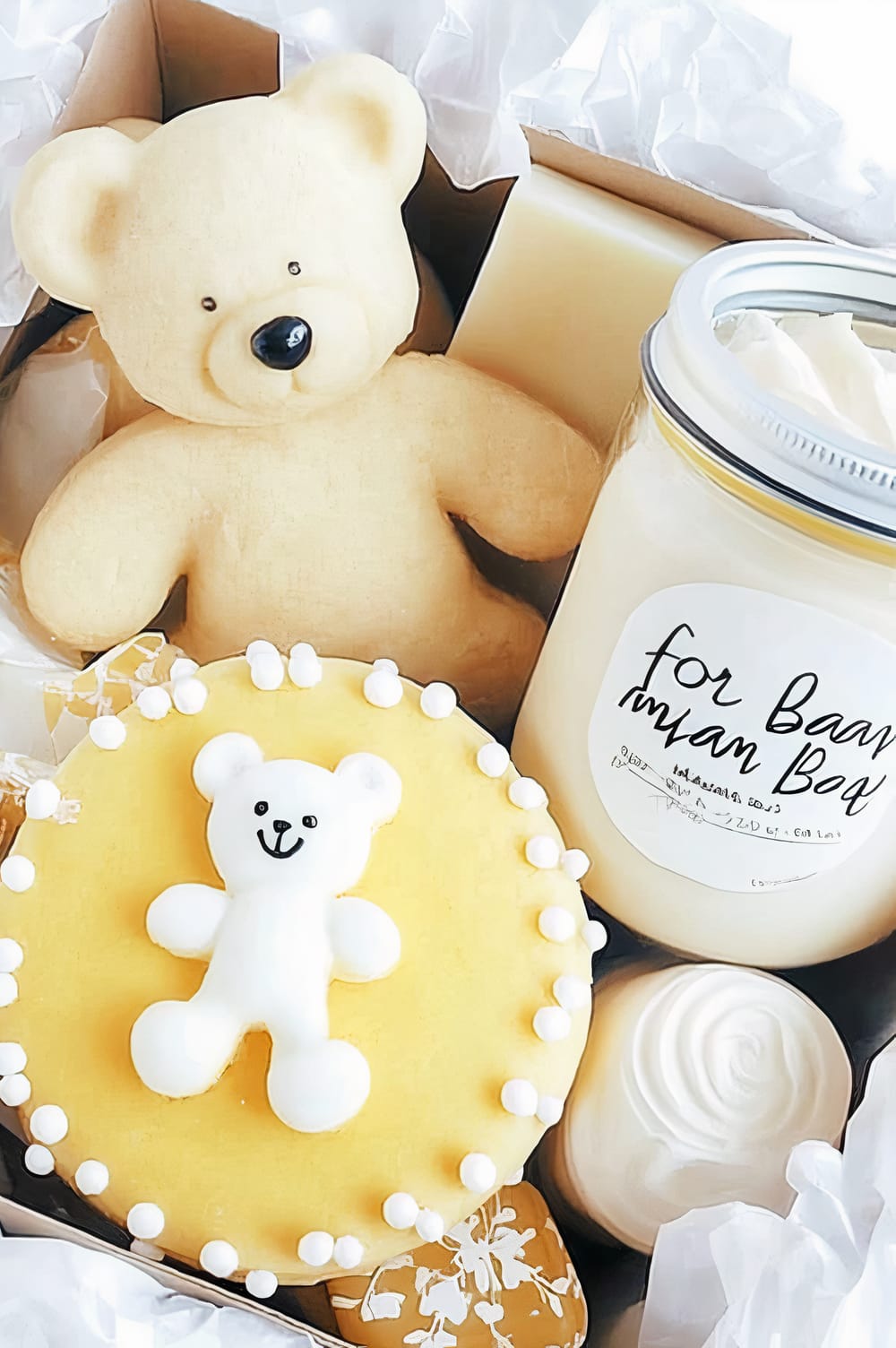 A gift box containing a plush teddy bear, a jar labeled "for Baby", and a yellow round cake decorated with a small white teddy bear-shaped icing topper and white icing dots. The items are nestled in white tissue paper.