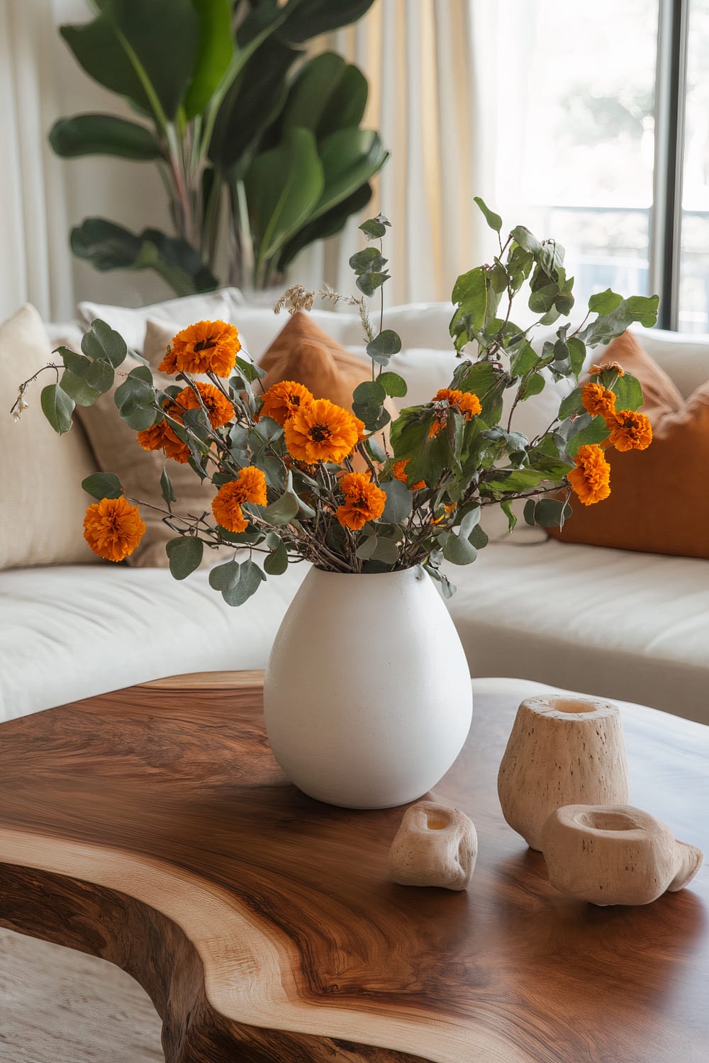 A nature-inspired Thanksgiving coffee table setup features a wooden table with a live edge. On the table, there is a simple white ceramic vase filled with dried eucalyptus and bright orange marigolds. Additionally, there are a few small wooden branches and natural stone candle holders, adding organic texture. Surrounding the table are earth-toned cushions on a linen sofa. In the background, large leafy plants and soft natural light filter through sheer curtains, creating a serene and uncluttered aesthetic.