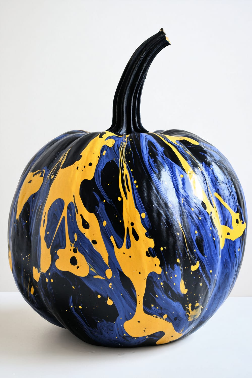 A close-up image of a pumpkin painted with a glossy black base. Over the black base, vibrant blue and yellow acrylic paint is splashed and marbled in an abstract pattern, creating a striking and dynamic visual effect. The pumpkin rests on a white surface against a plain white background.