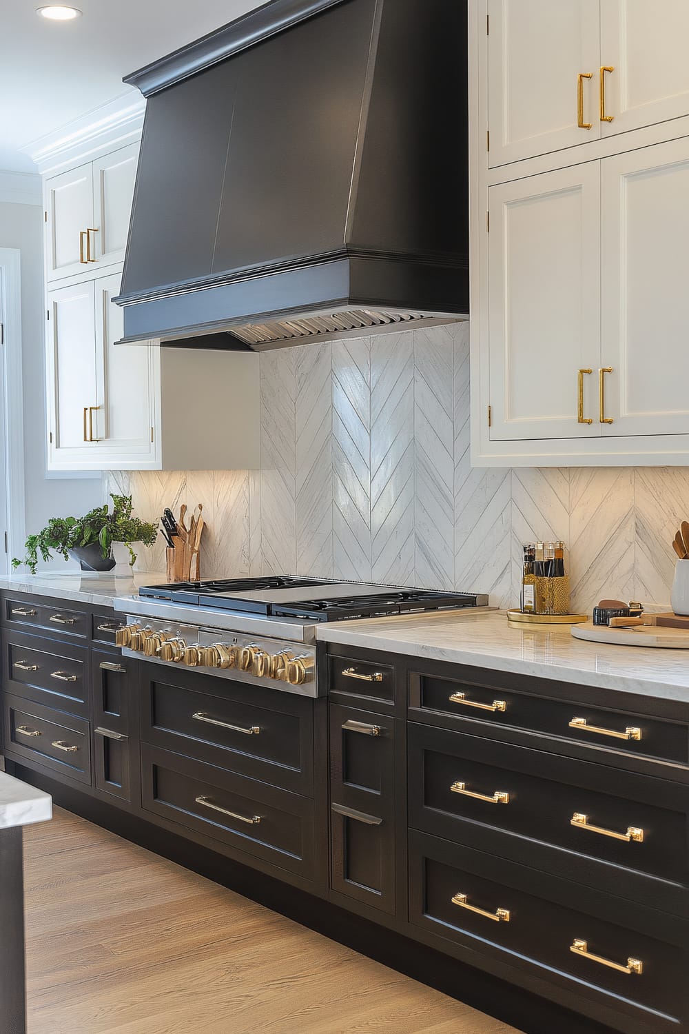 A modern kitchen with sophisticated black lower cabinetry, white upper cabinets, and a striking black range hood. The black cabinets are equipped with brass handles, and the backsplash features a herringbone pattern in white marble. The countertops are made of white marble, and the kitchen floor is wooden. The setup includes a stainless-steel range with brass knobs and a variety of kitchen tools and condiments strategically placed on the countertops.