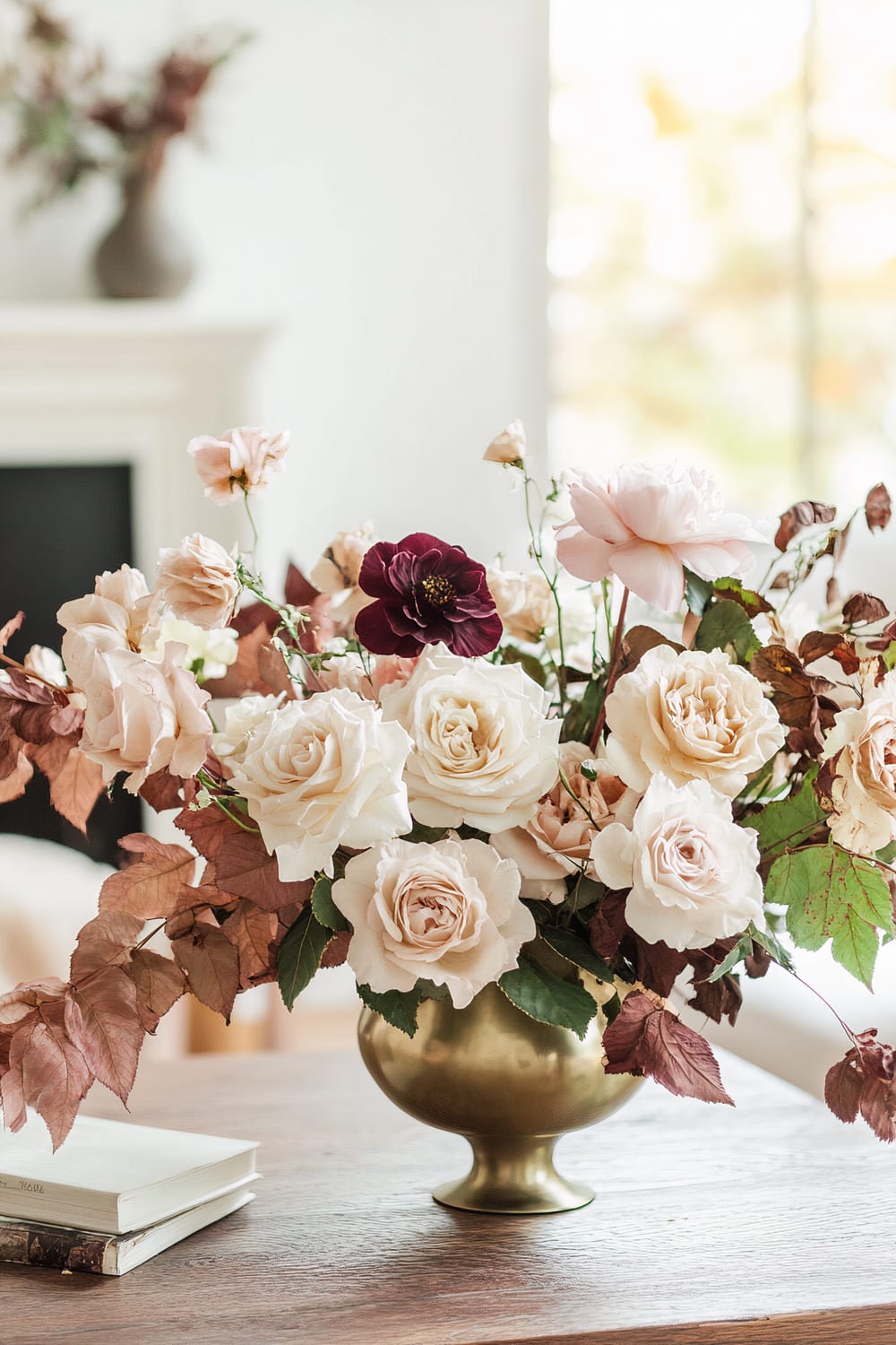 A floral arrangement in a brass vase placed on a wooden table. The bouquet includes white and light pink roses, a single dark maroon flower, and some dried leaves in shades of brown. In the background, a blurred vase with additional flowers and foliage sits on a mantle or shelf, with light filtering through a nearby window.