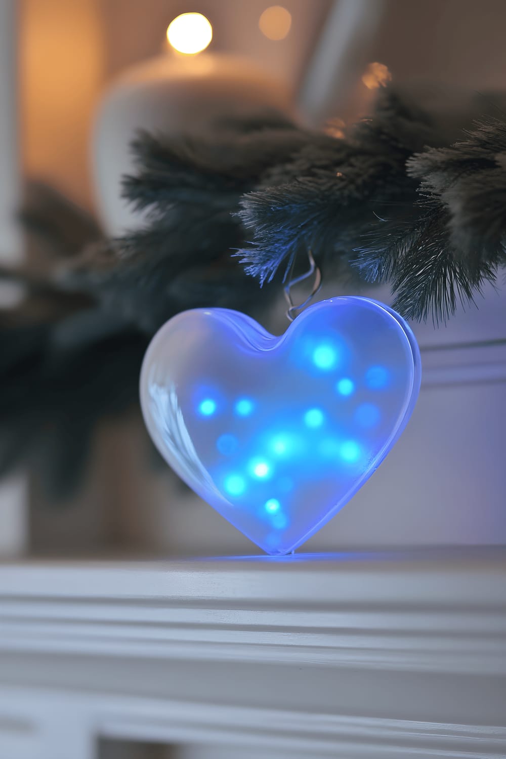 A frosted glass heart-shaped ornament containing small floating blue LED lights sits on a white mantel. The lights inside the ornament illuminate softly, casting a serene blue glow that creates a beautiful contrast against the neutral white backdrop.