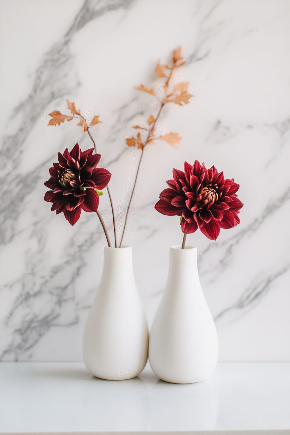 Two white ceramic vases with minimalist design, each holding a single stem of dark red dahlia flowers, are placed on a clean marble surface against a white marble background with grey veins.