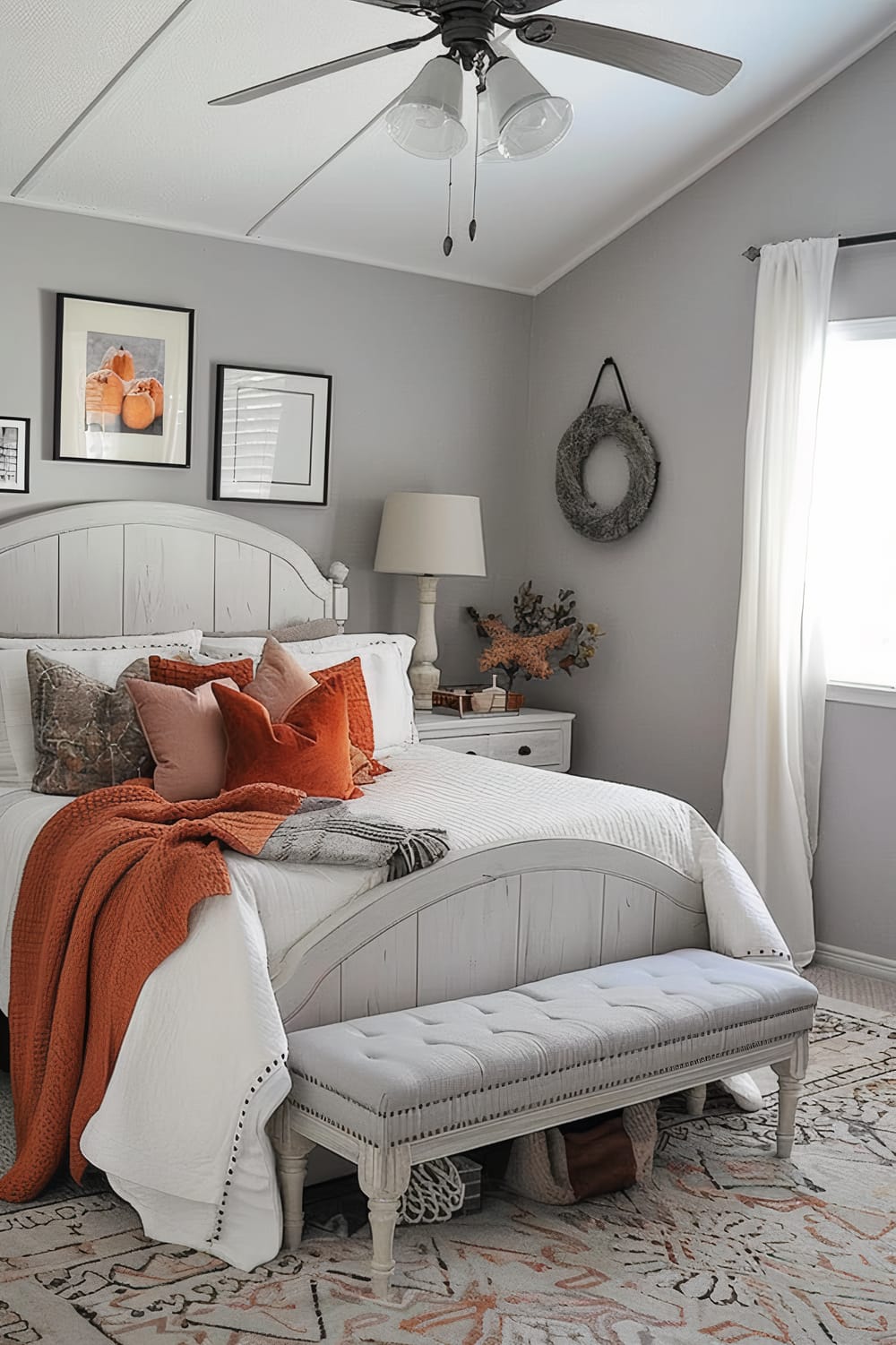 An elegantly designed bedroom featuring a white wooden bed adorned with white bedding and multiple pillows in orange and neutral tones. A coordinating orange blanket is draped at the foot of the bed, complemented by a beige tufted bench. The walls are painted light gray and decorated with minimalist framed art and a decorative wreath. A white nightstand supports a classic table lamp, while a window with white curtains allows natural light to brighten the space. A patterned area rug and ceiling fan complete the room.