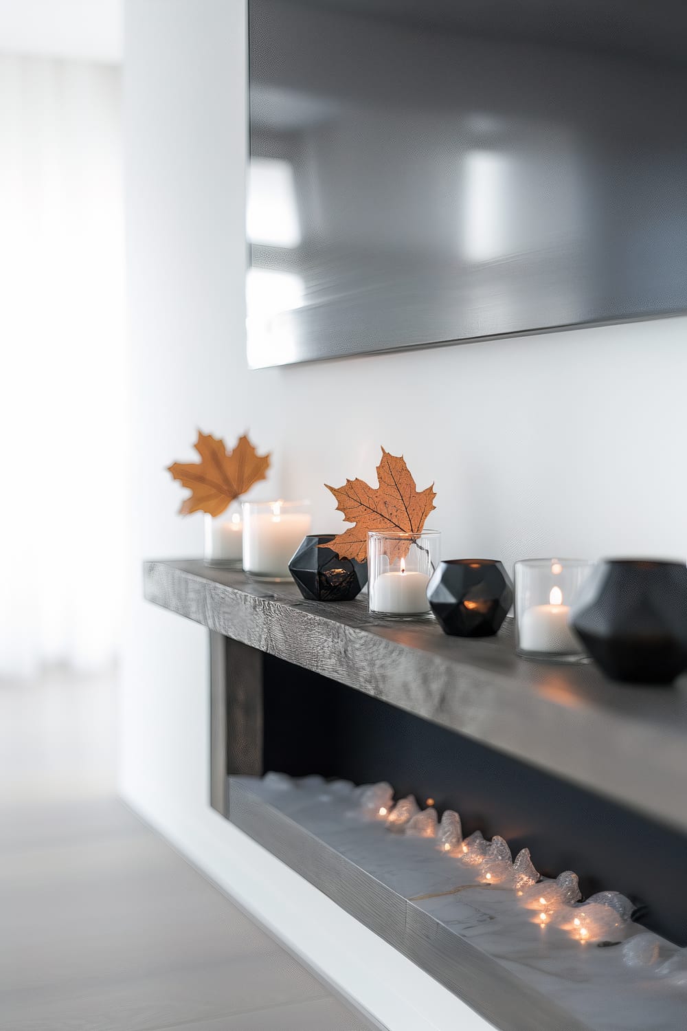 An elegant modern fireplace mantle adorned with various sleek black and clear candle holders containing lit white candles. Decorative autumn leaves are used as accents inside some of the candle holders. There is an artificial fire element in the fireplace simulating glowing embers. Above the mantle, a large, flat-panel TV is mounted on the wall. The setting is minimalistic with neutral tones and subtle lighting.