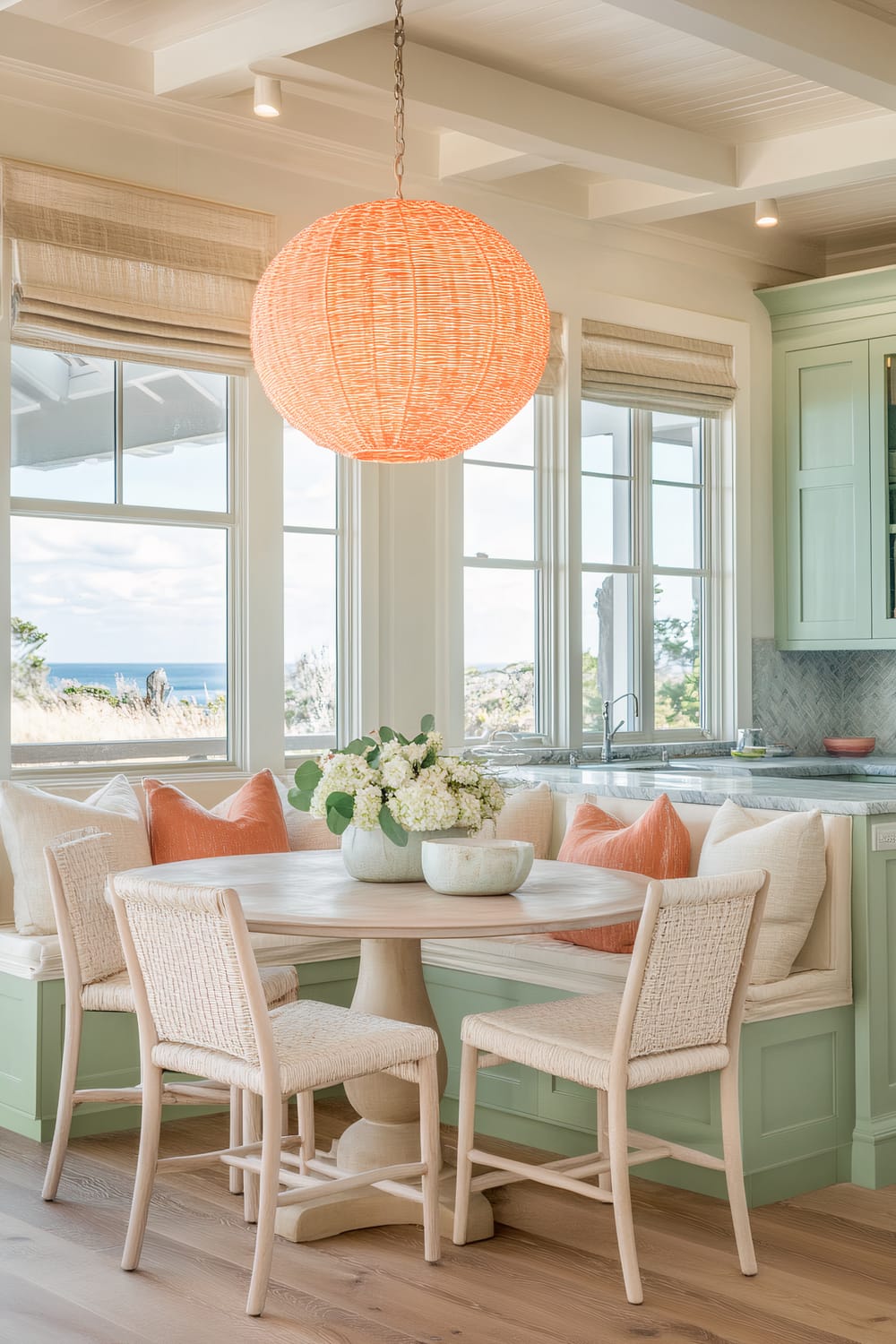 A light and airy kitchen dining area featuring a round wooden table with four woven chairs. The space has a large, bright orange woven pendant light hanging from the ceiling. A built-in bench with mint green cabinetry beneath and adorned with various throw pillows, including bright orange and neutral tones, surrounds the table. The large windows offer an ocean view, contributing to the coastal vibe.