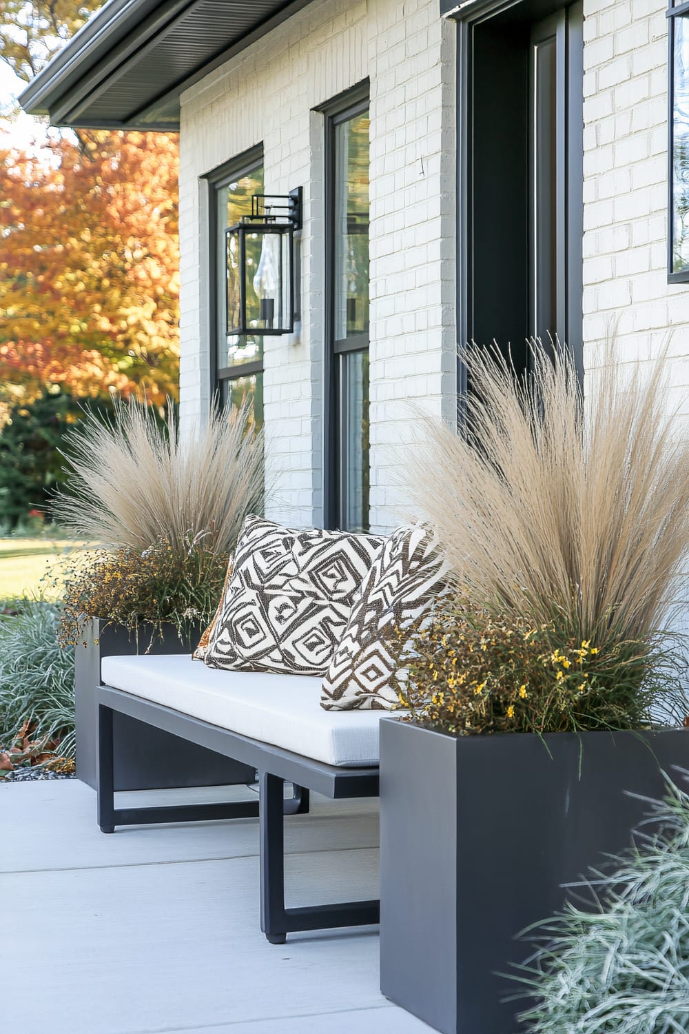 An outdoor modern seating area is depicted with a sleek black frame bench featuring plush white cushions and two geometric-patterned throw pillows in black and white. The bench is flanked by large, black cubic planters filled with ornamental grasses and small flowering plants. The backdrop includes a white brick exterior with black-framed windows and a black lantern-style outdoor wall sconce.