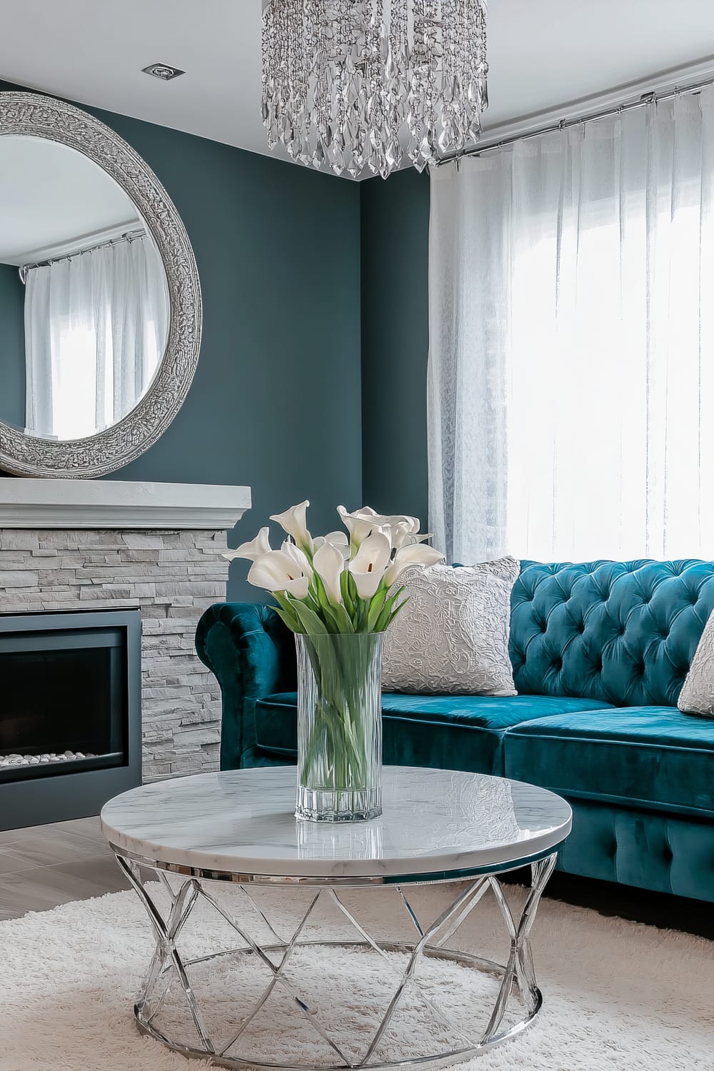 A Parisian-inspired living room featuring a teal tufted velvet sofa with white lace throw pillows. A round marble coffee table with polished chrome legs sits on a cream rug. A large silver-framed mirror hangs above a modern stone fireplace. Sheer white curtains adorn light teal walls, and a vase of white lilies sits on the coffee table.