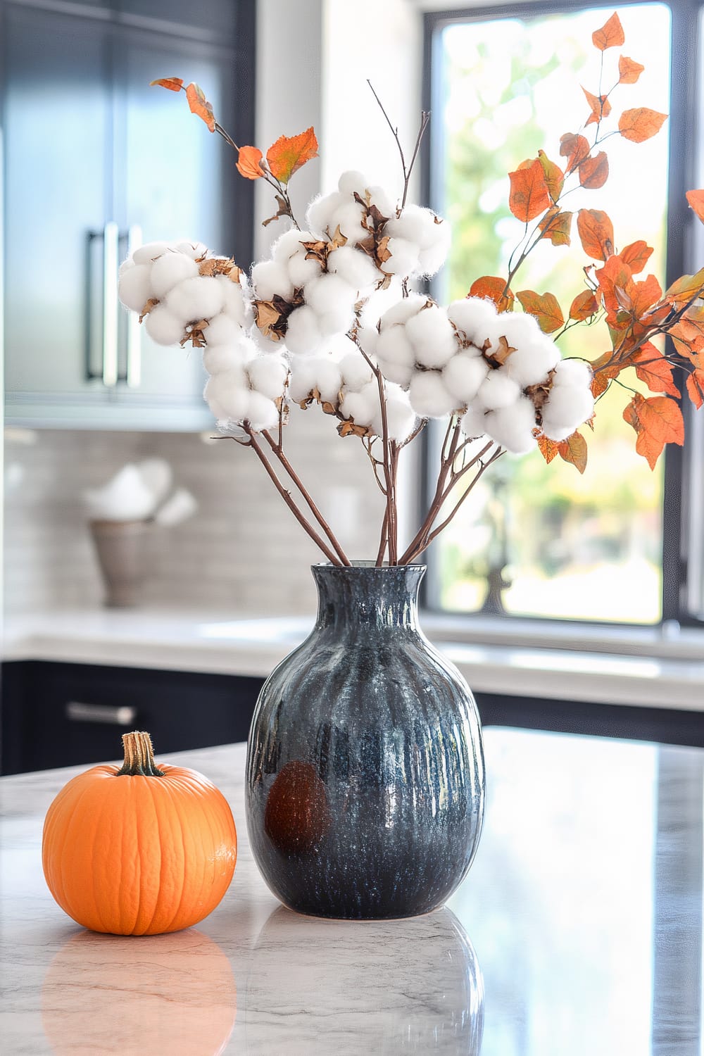 An elegant kitchen countertop features a dark, glossy vase with branches of white cotton flowers and orange autumn leaves arranged in it. Beside the vase sits a small, vibrant orange pumpkin. The background includes a sleek kitchen with dark cabinets, a light tiled backsplash, and large windows allowing natural light to flood the space.