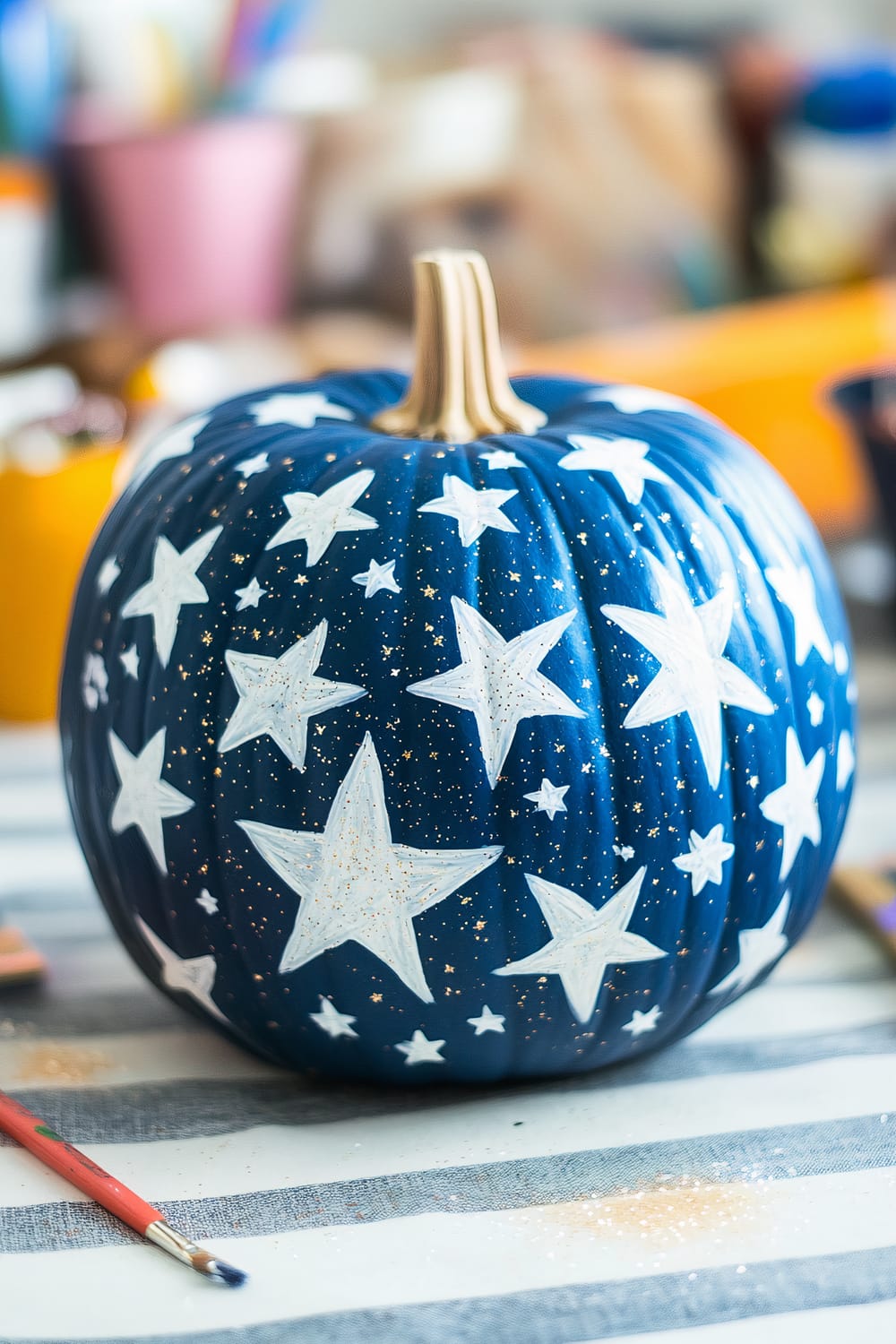 A blue pumpkin with white stars painted on its surface, resembling a night sky. The background is slightly blurred, showing art supplies such as paintbrushes and containers.