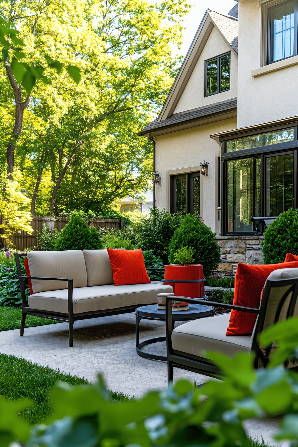 A well-maintained backyard featuring beige outdoor furniture with red and orange cushions on a clean, simple concrete patio. It’s bordered by verdant bushes and a manicured lawn. A decorative vibrant potted plant is positioned on the patio adding color to the scene. The house facade visible in the background has large windows, reinforcing the indoor and outdoor connection.