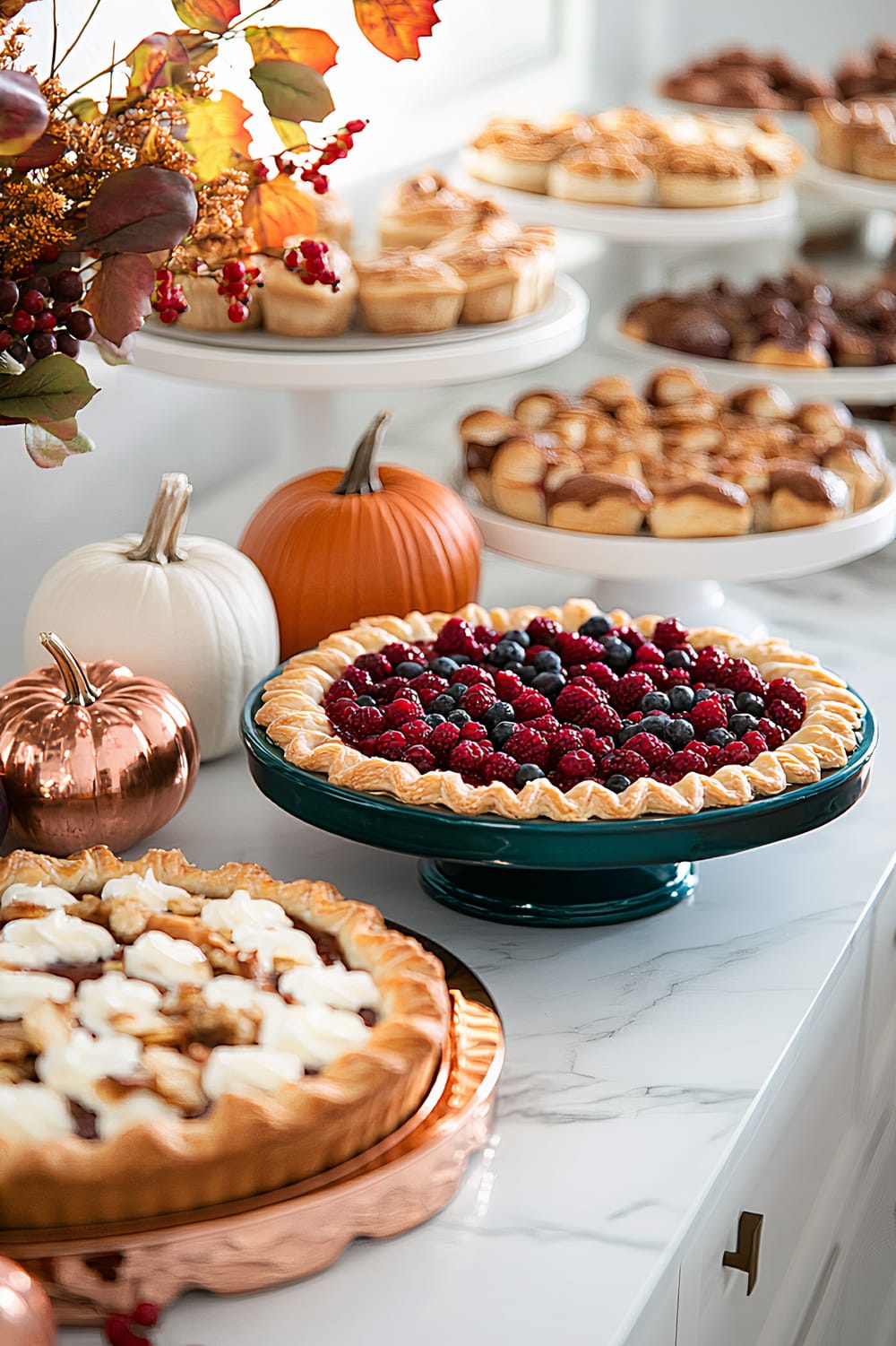 An elegant dessert table set for Thanksgiving with a sleek steel table and white marble countertops. It features a variety of gourmet pastries and pies, including a berry-topped pie on a teal stand and a cream-topped pie on a copper tray. The table is decorated with teal and copper serving trays, minimalist decorative pumpkins, and autumn branches. Soft ambient lighting enhances the warm and inviting display.