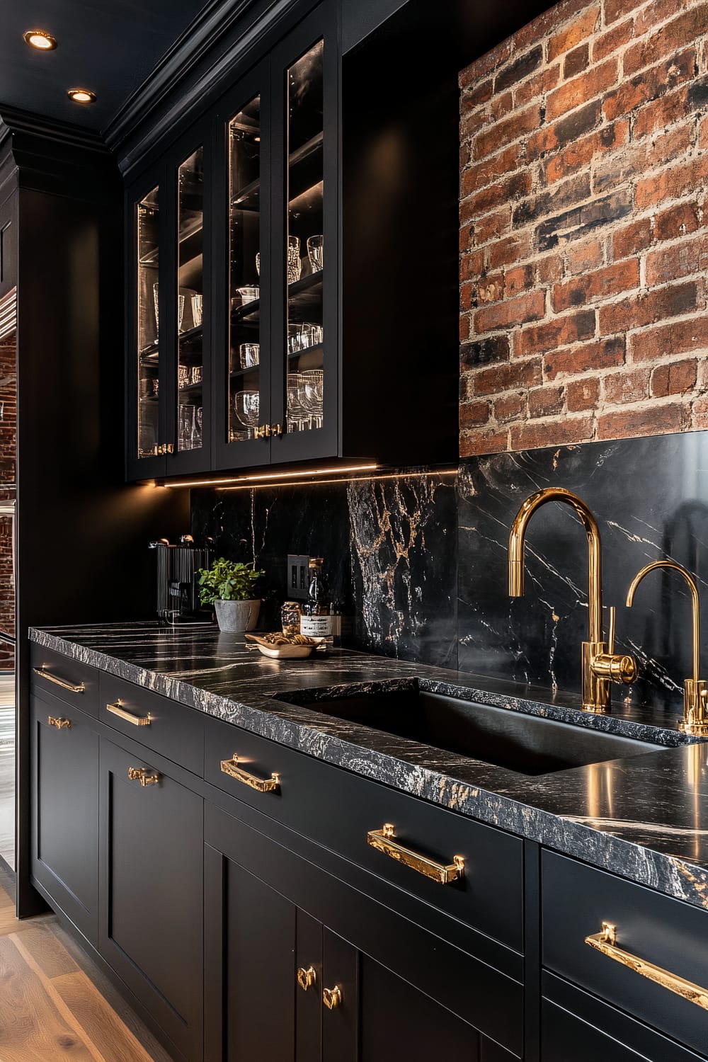 A sophisticated, modern kitchen features striking black cabinetry with minimalistic gold handles. The space is highlighted by a luxurious black marble countertop and matching backsplash, both showcasing white and gold veins. Open shelving above displays an array of glassware while the exposed brick wall adds a rustic touch. A sleek gold faucet and under-cabinet lighting provide additional elegance and warmth to the space.