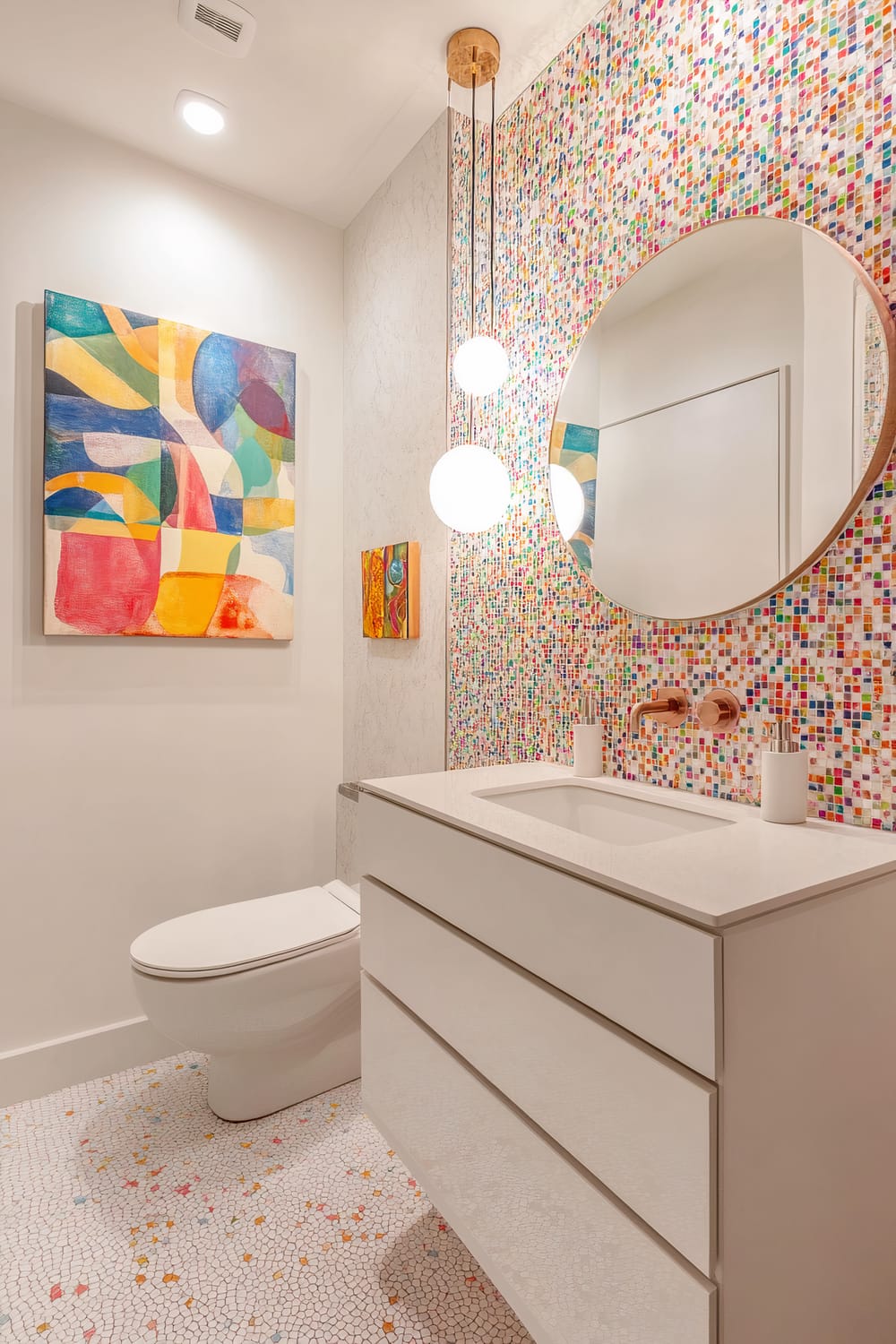 A bathroom featuring a white toilet, a vanity with smooth white drawers, a rectangular sink, and a marble countertop. Above the vanity hangs a circular mirror with a wooden frame, reflecting the room's colorful decor. The back wall is adorned with multicolored mosaic tiles, creating a vibrant backdrop. Pendant lights with round white globes hang from the ceiling. On the wall to the left of the vanity, a large abstract painting with bold, colorful shapes complements a smaller, similar artwork below it. The floor is decorated with small, white hexagonal tiles interspersed with multicolored accents.