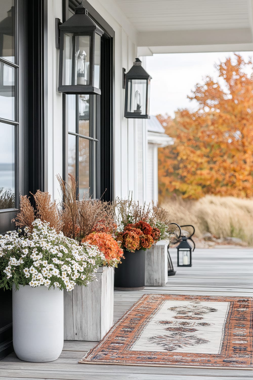 An outdoor porch area featuring a set of black-framed windows and door with black lantern-style sconces affixed to the wall. The porch has potted plants with flowers in various colors placed on the wooden deck. A traditional-patterned area rug with an intricate border is spread across the floor, and the background showcases autumn trees with orange, brown, and yellow leaves.