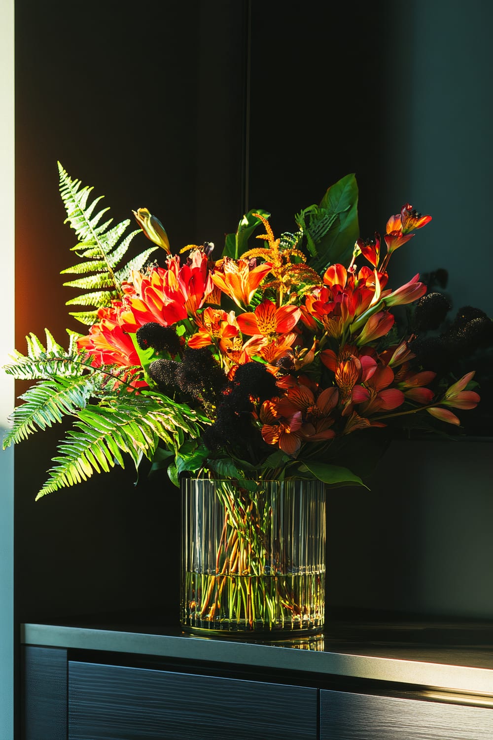 A vibrant floral arrangement in a transparent cylindrical vase is placed on a dark-colored modern furniture unit. The bouquet features predominantly orange and red flowers, accented with green ferns and some dark-colored floral elements. The arrangement is illuminated by warm sunlight coming from the left side, casting a soft glow over the scene.