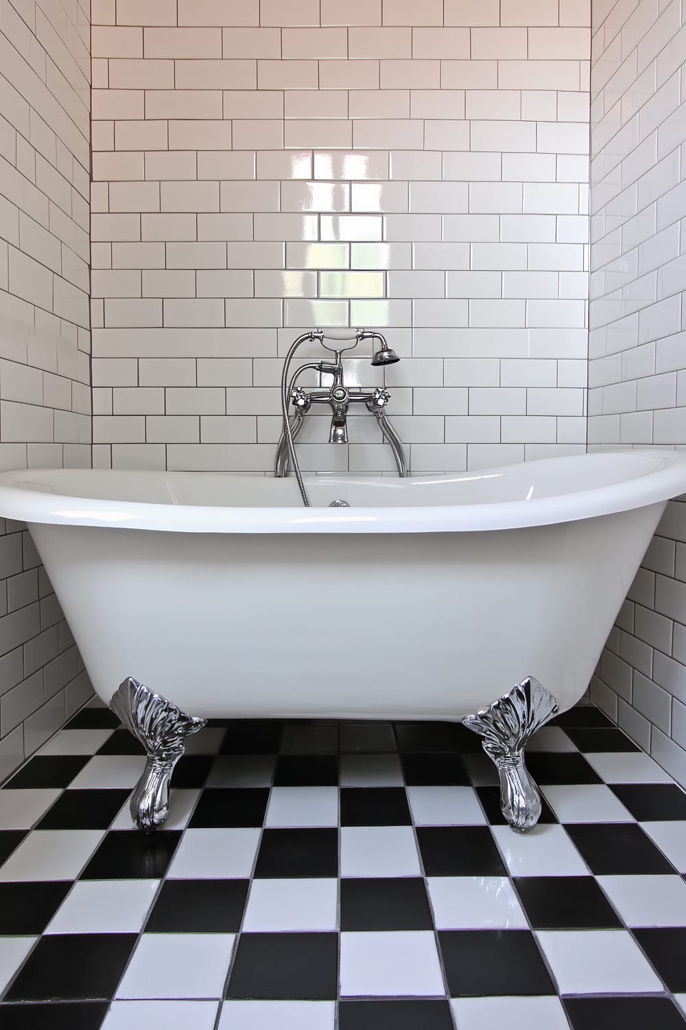 A bright, white bathroom with a freestanding clawfoot bathtub. The walls are adorned with glossy white subway tiles, and the floor features a striking black and white checkerboard pattern. The bathtub has traditional chrome claw feet and an elegant, vintage-style faucet.