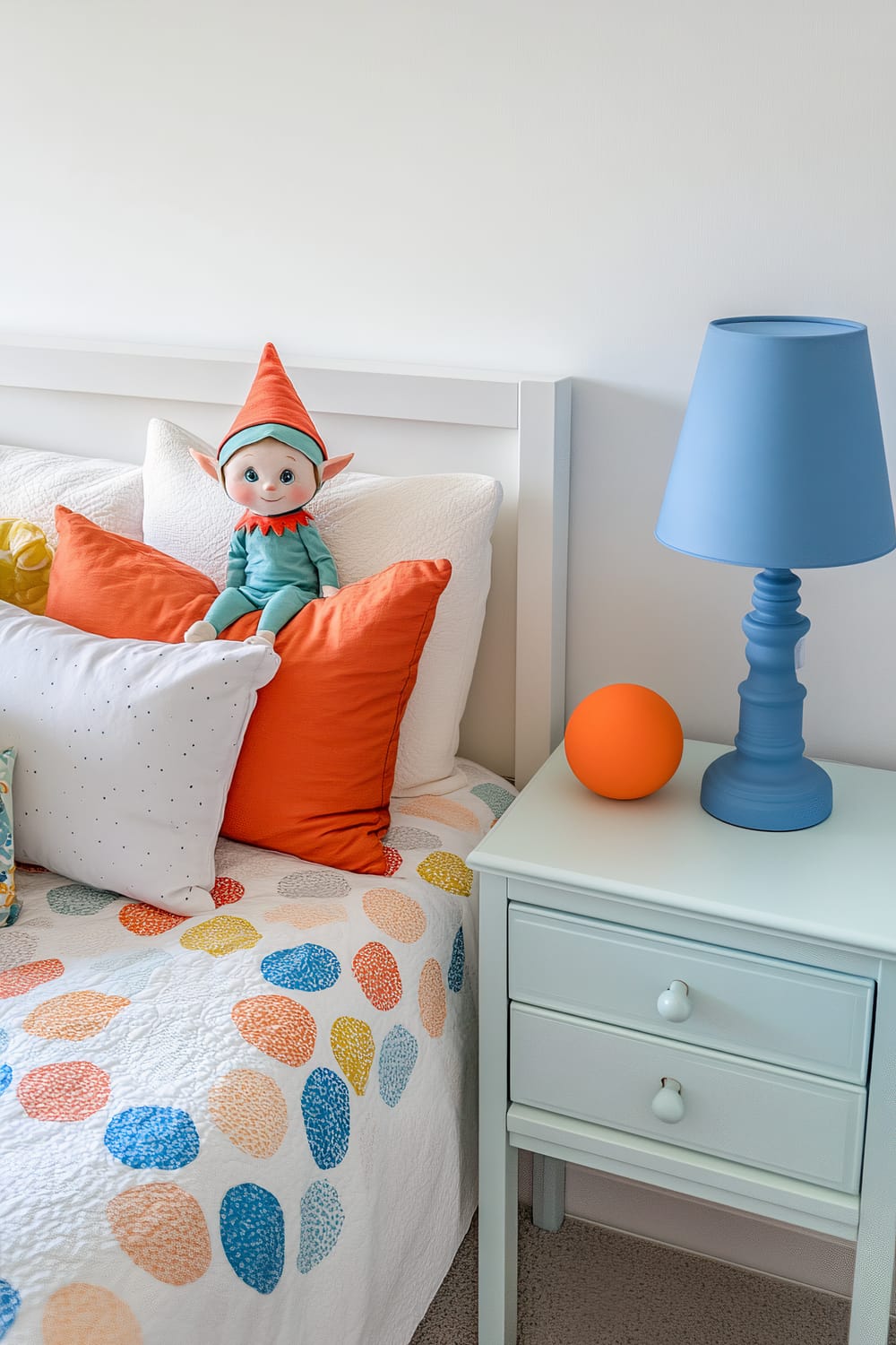 A charming bedroom scene featuring a whimsical elf doll seated among pillows on a white bed, a blue nightstand with a matching blue lamp, and an orange ball.