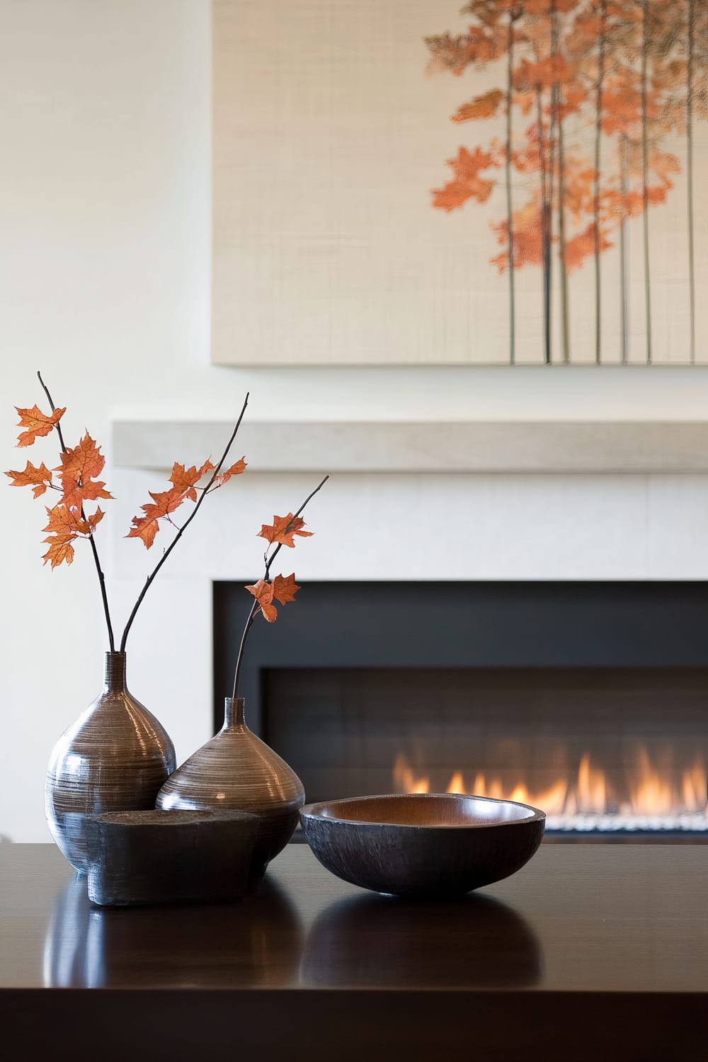 A close-up of a sophisticated living room setting featuring a modern fireplace with an abstract painting of autumn leaves hanging above it. Two ceramic vases with branches of red and orange leaves and a dark bowl are elegantly placed on a dark wooden table in the foreground.