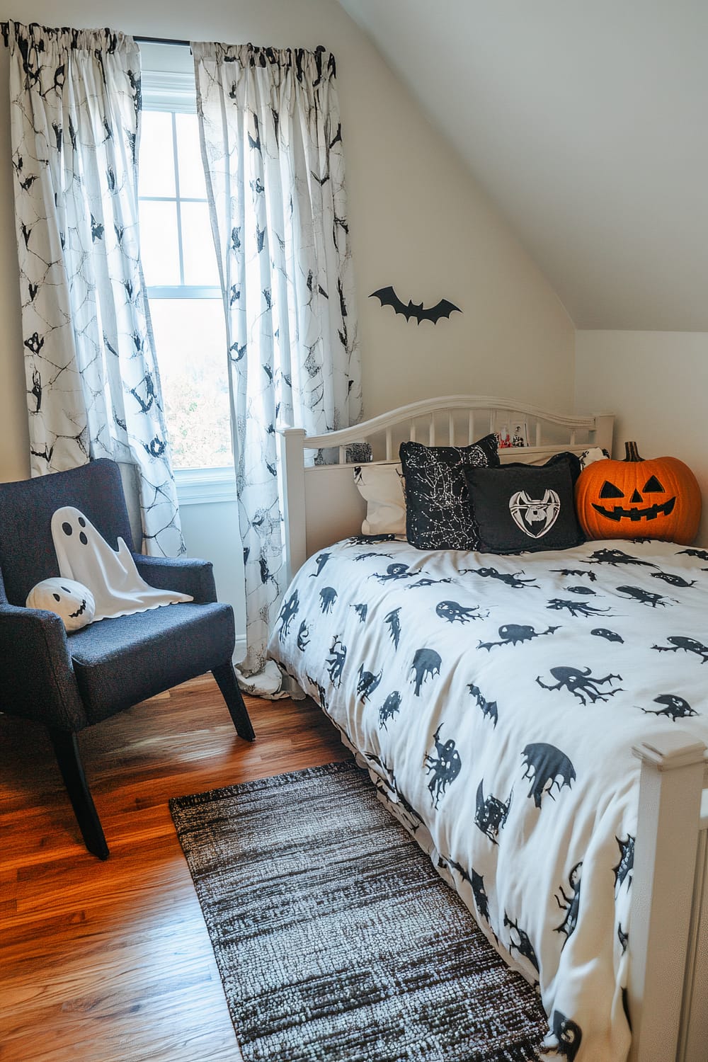 A small bedroom is decorated with a Halloween theme. The bed has white bedding with black spooky creature designs. There are black and white pillows, including one with a bat motif. A large carved pumpkin with a jack-o'-lantern face sits on the bed. The window is adorned with white curtains featuring black spider web patterns. A dark gray chair has a ghost-shaped cushion and a jack-o'-lantern pillow. A bat decoration is mounted on the wall.