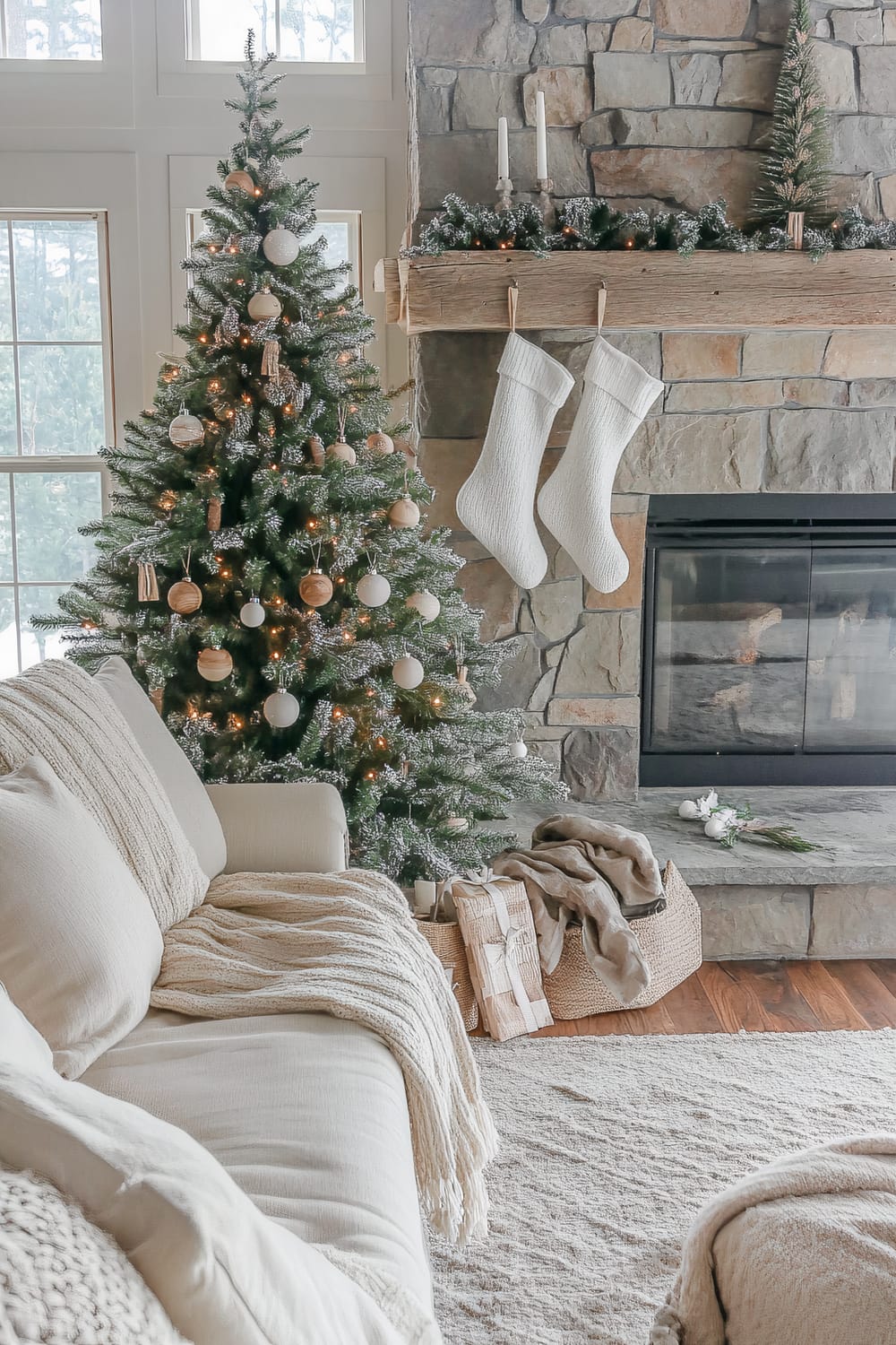 An inviting living room decorated for the Christmas season. The room features a tall, ornamented Christmas tree adorned with white and wooden baubles. Beside the tree is a stone fireplace, decorated with green garland, white stockings, and white candles on the mantel. A beige sofa with cream-colored throws and pillows sits in the foreground, and a basket with wrapped presents and a cozy blanket is placed nearby.