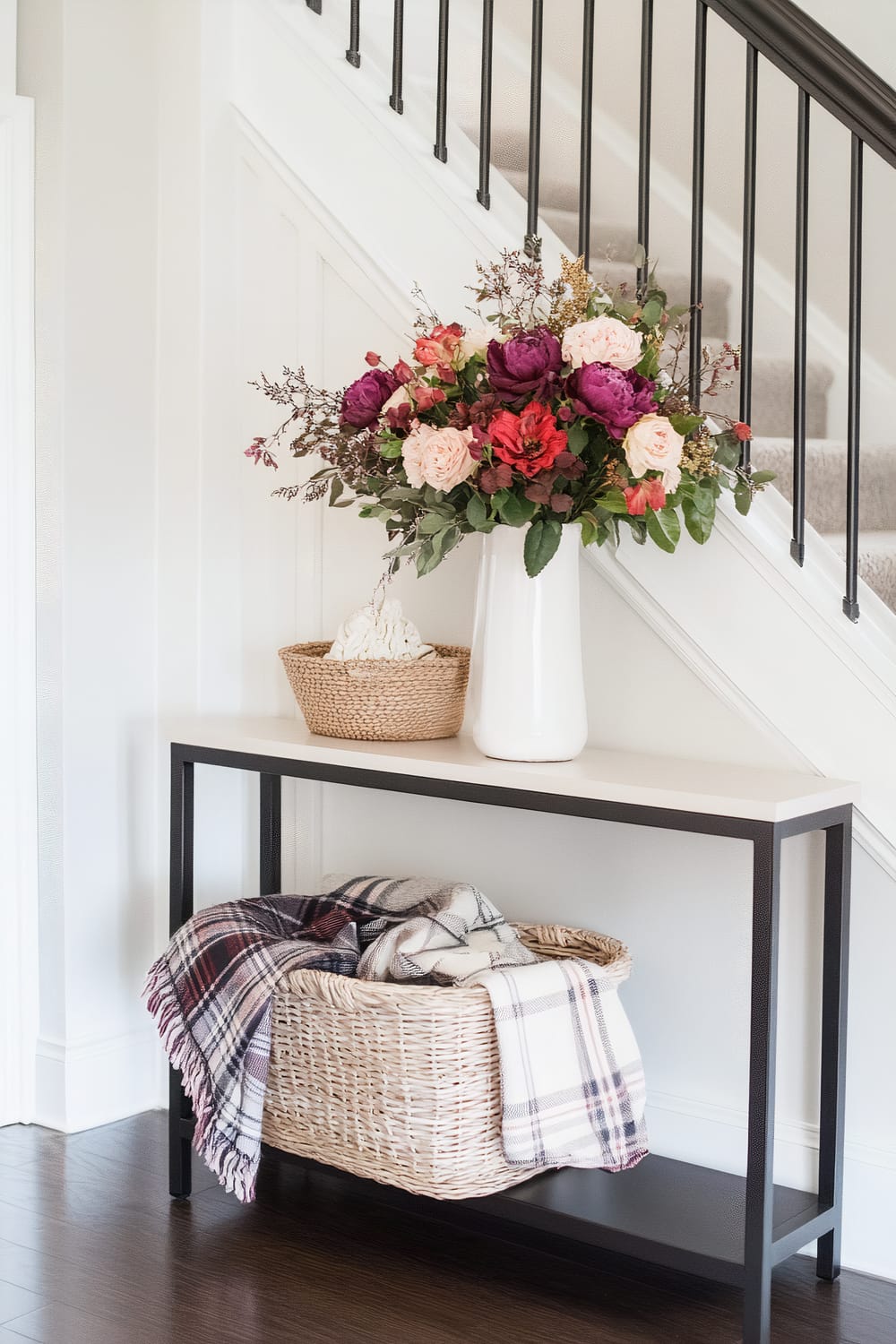 A modern entryway features a black metal and white surface console table against a white wall beneath a staircase with black railings. On the table is a tall white vase with a vibrant floral arrangement of purple, red, and pink flowers, and a small woven basket. Below the console table is a wicker basket holding a plaid blanket.