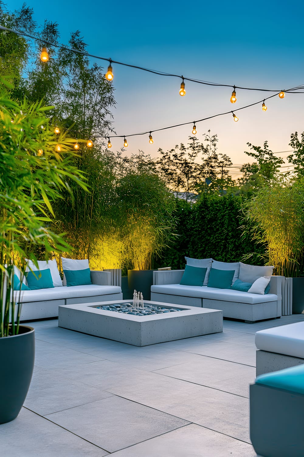 A serene modern garden patio during twilight features a sleek grey concrete platform, minimalist white outdoor furniture with plush teal cushions, a central low-maintenance stone water fountain, vibrant green bamboo plants in black ceramic pots, and string lights overhead creating a warm glow.