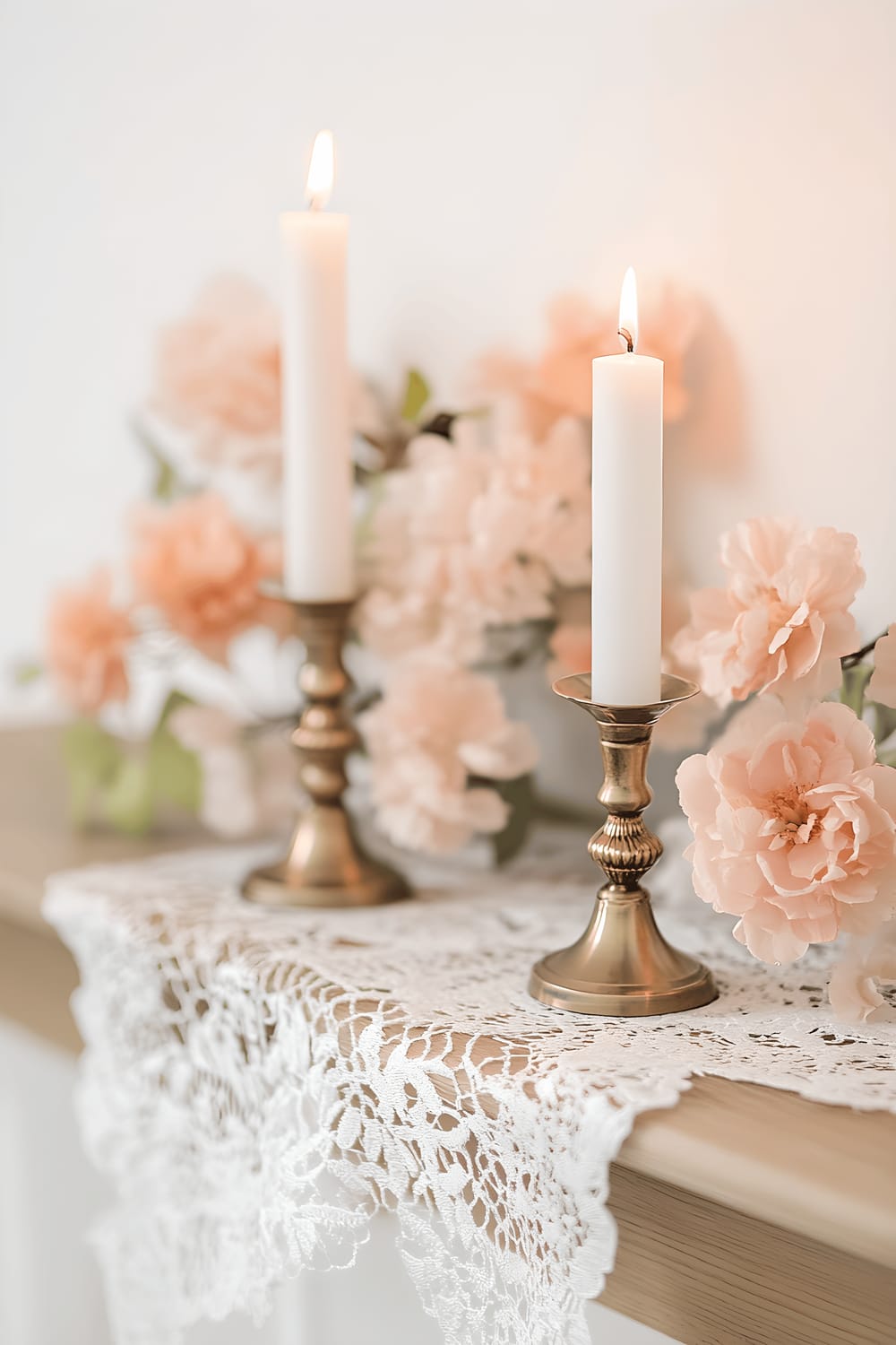 An image of a delicate white lace table runner spread across a light oak mantel. The mantel is graced with two brass candle holders, each holding a lit white candle. Soft pink faux florals are beautifully scattered around the candles, all illuminated gently by the candlelight.